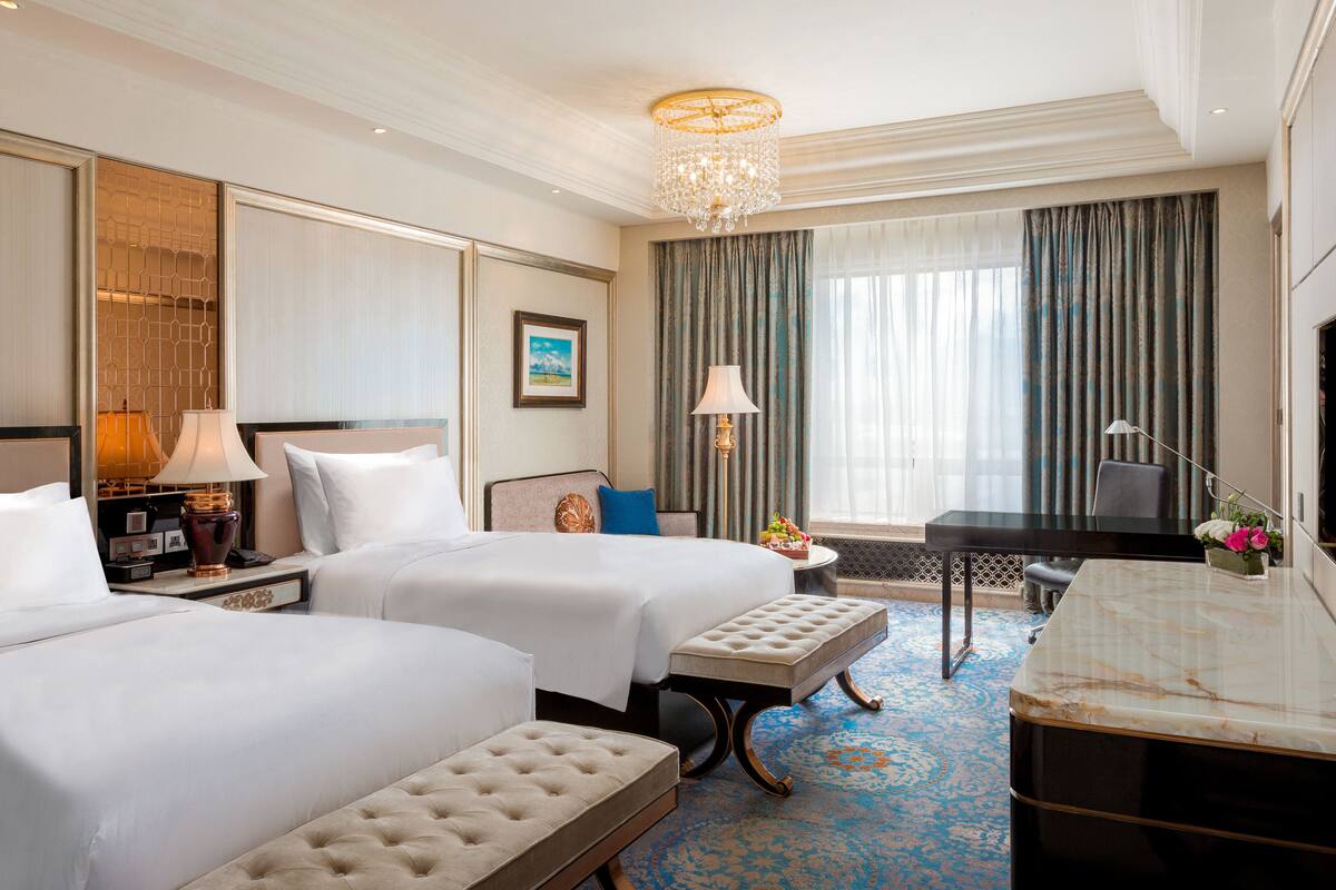 Quarto de hotel com duas camas, uma escrivaninha, uma cadeira, uma televisão e um candelabro.
