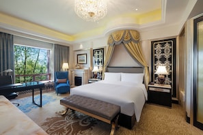 Habitación de hotel con una cama grande, una zona de estar con una silla y una mesa, una chimenea y un balcón con vistas a árboles.