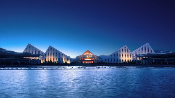 Un hotel moderno con un edificio central y tres extensiones simétricas, todas con un diseño geométrico, situado junto a una masa de agua.