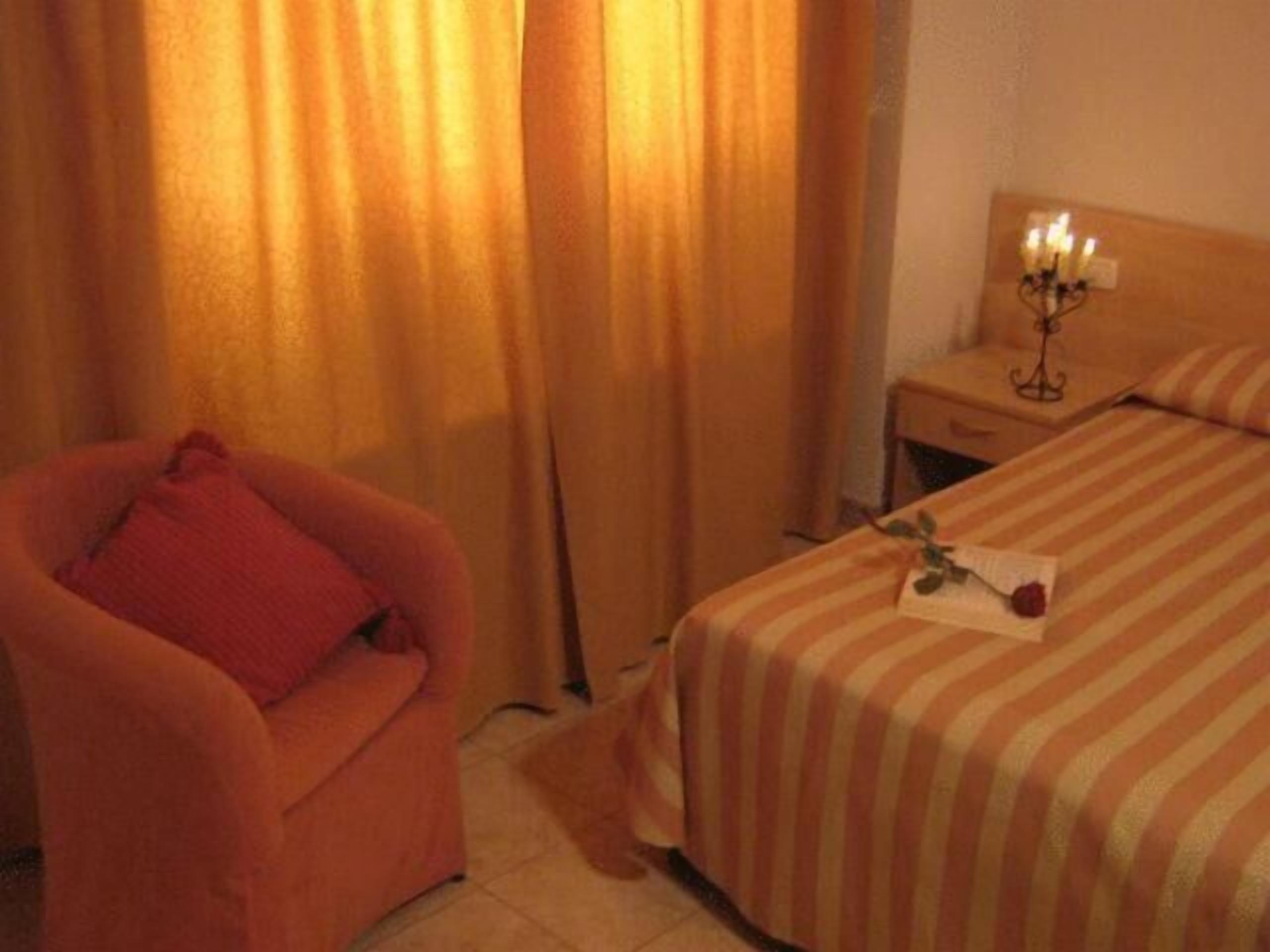 Una habitación de hotel con una cama a rayas, una silla con un cojín rojo y una mesita de noche con un candelabro.