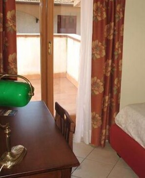 Een kamer met een houten tafel, een groene tas, een bed met een rode spreidek en uitzicht op een balkon door een deur.