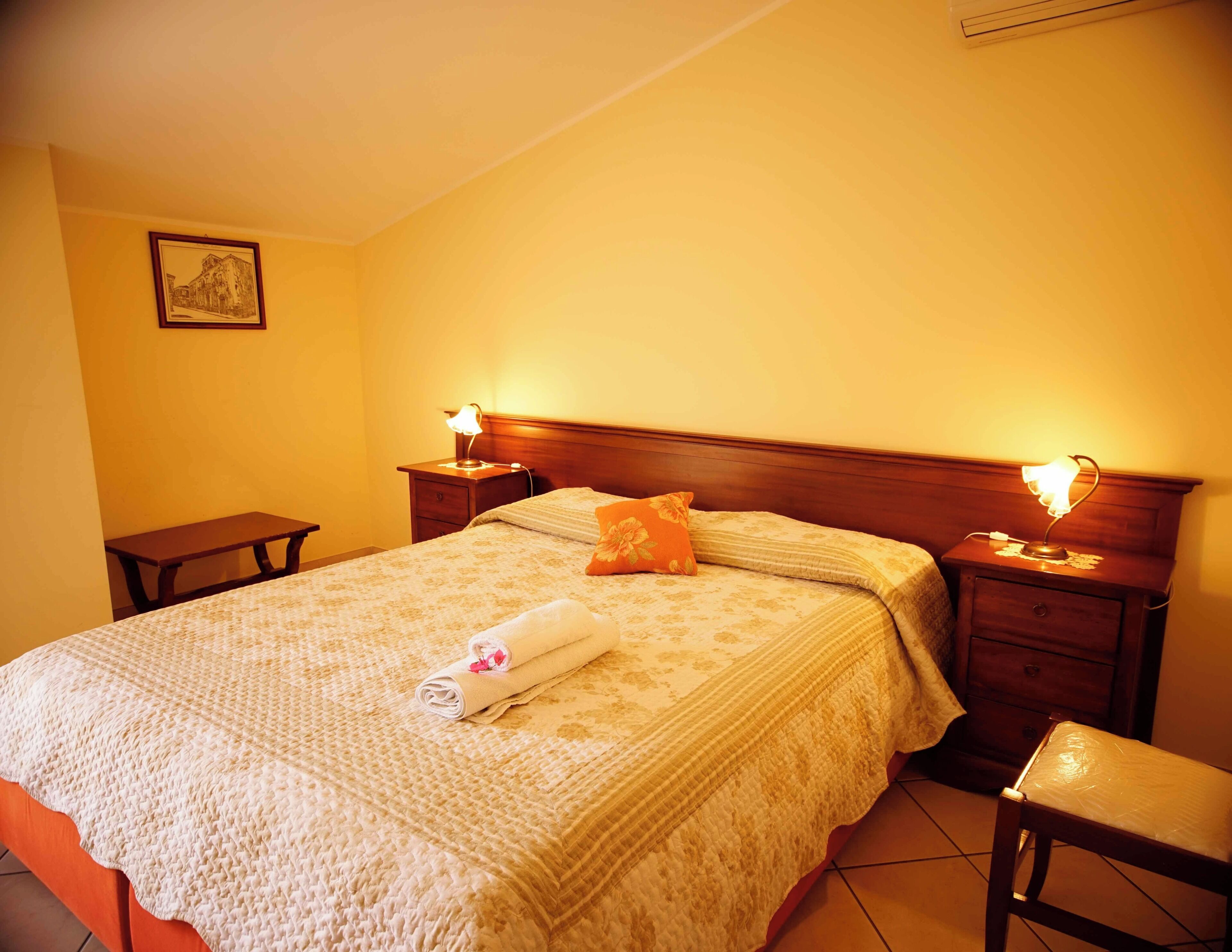 A neatly made bed with a rolled towel, two bedside tables with lamps, and a small table in a hotel room.