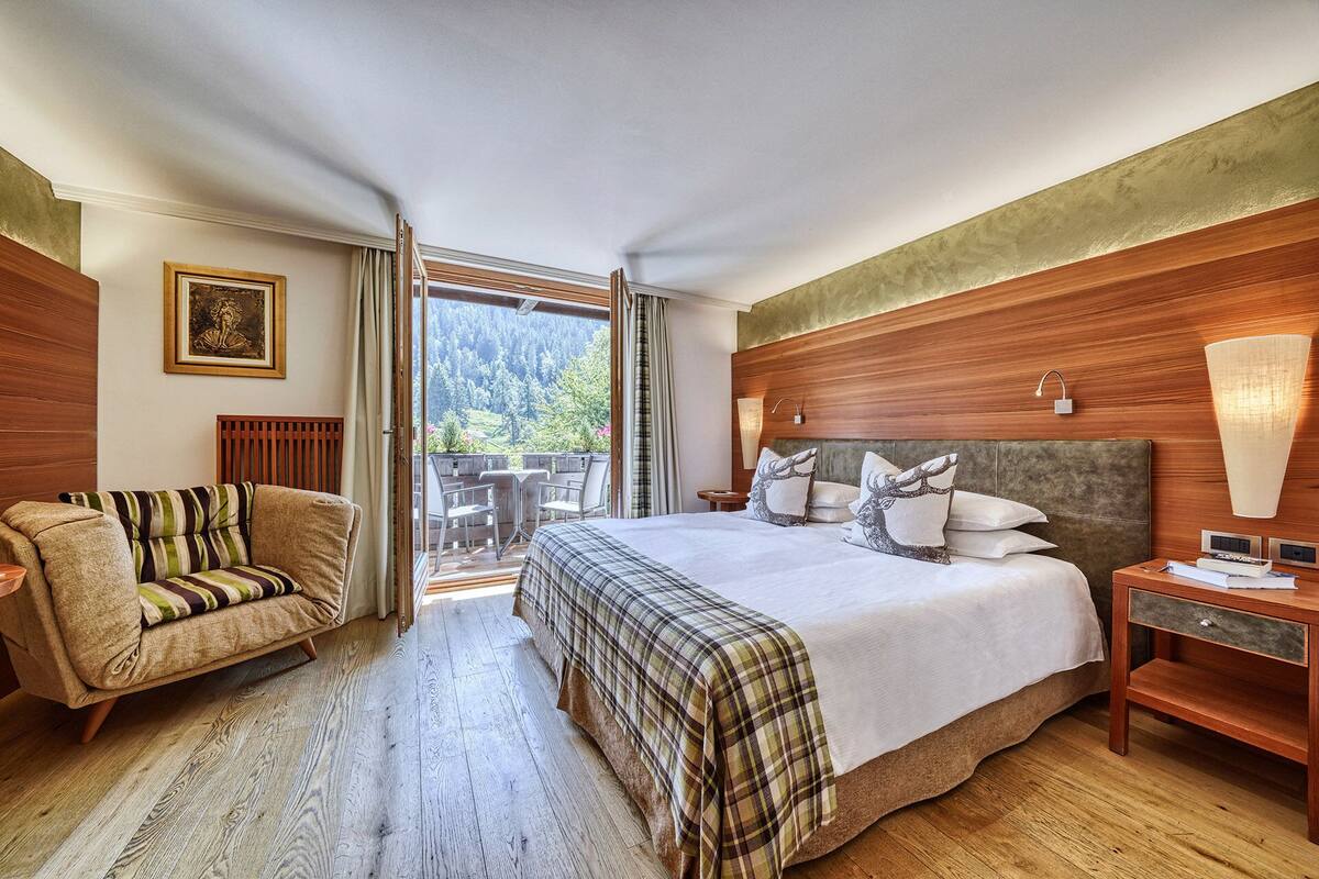 Een hotelkamer met een groot bed, een bank, een houten vloer, een balkon met uitzicht op bomen en een schilderij aan de muur.