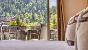 Una habitación de hotel con una cama, un balcón de madera y vistas a una montaña boscosa y edificios.