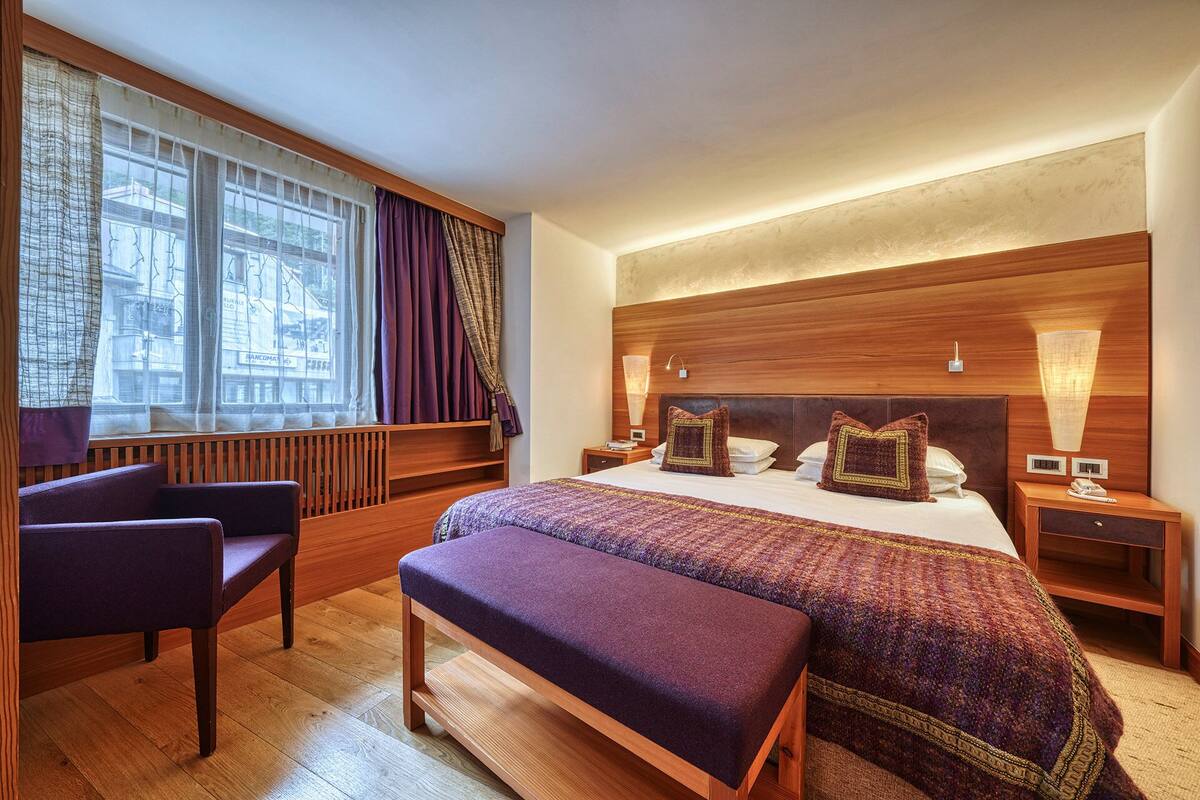 Hotelkamer met een groot bed, houten hoofdeinde, bedlampen, een bankje en uitzicht op de stad door het raam.