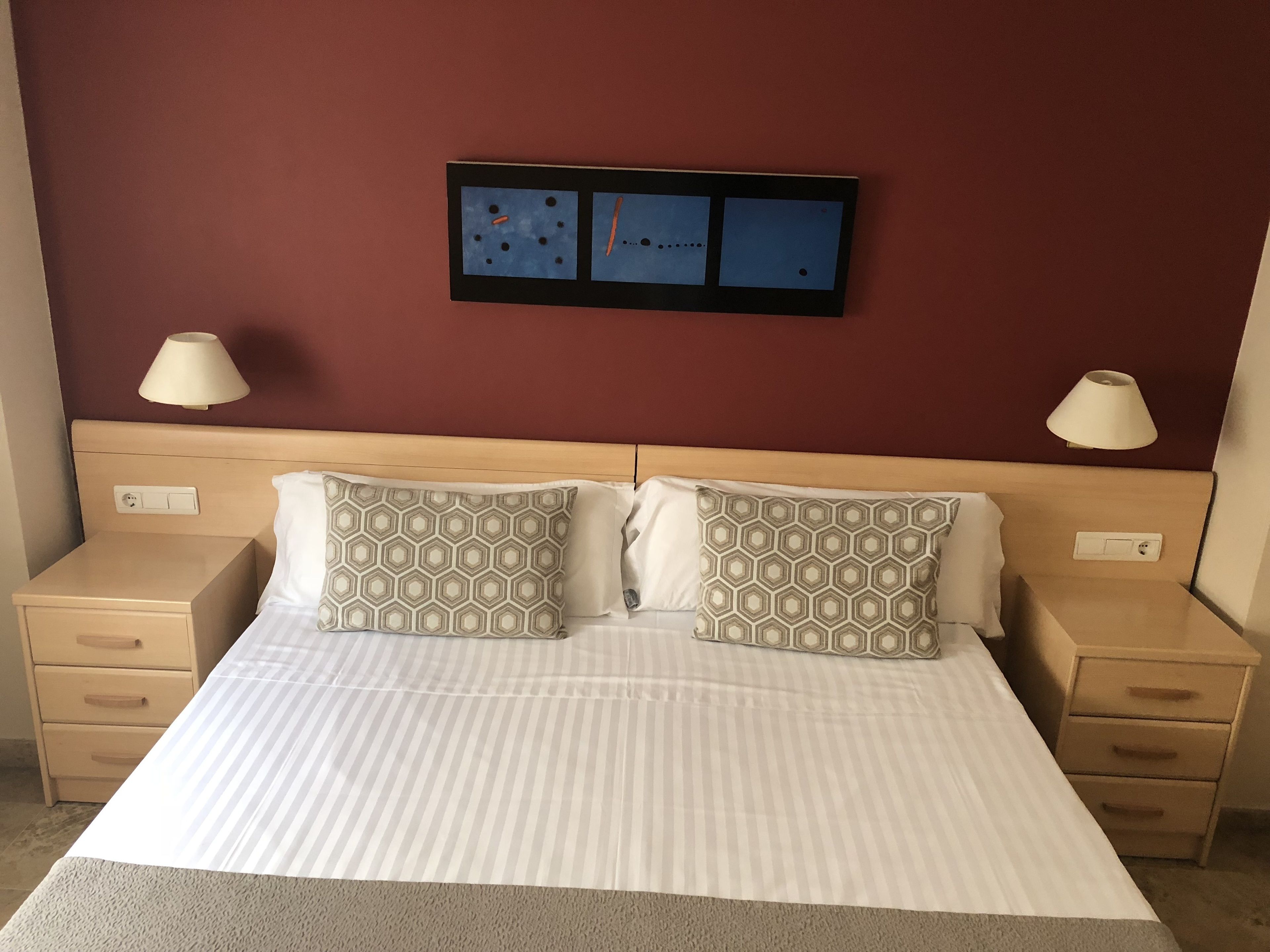 Una cama bien hecha con almohadones estampados, un cabecero, mesitas de noche con lámparas y un cuadro colgado en la pared.