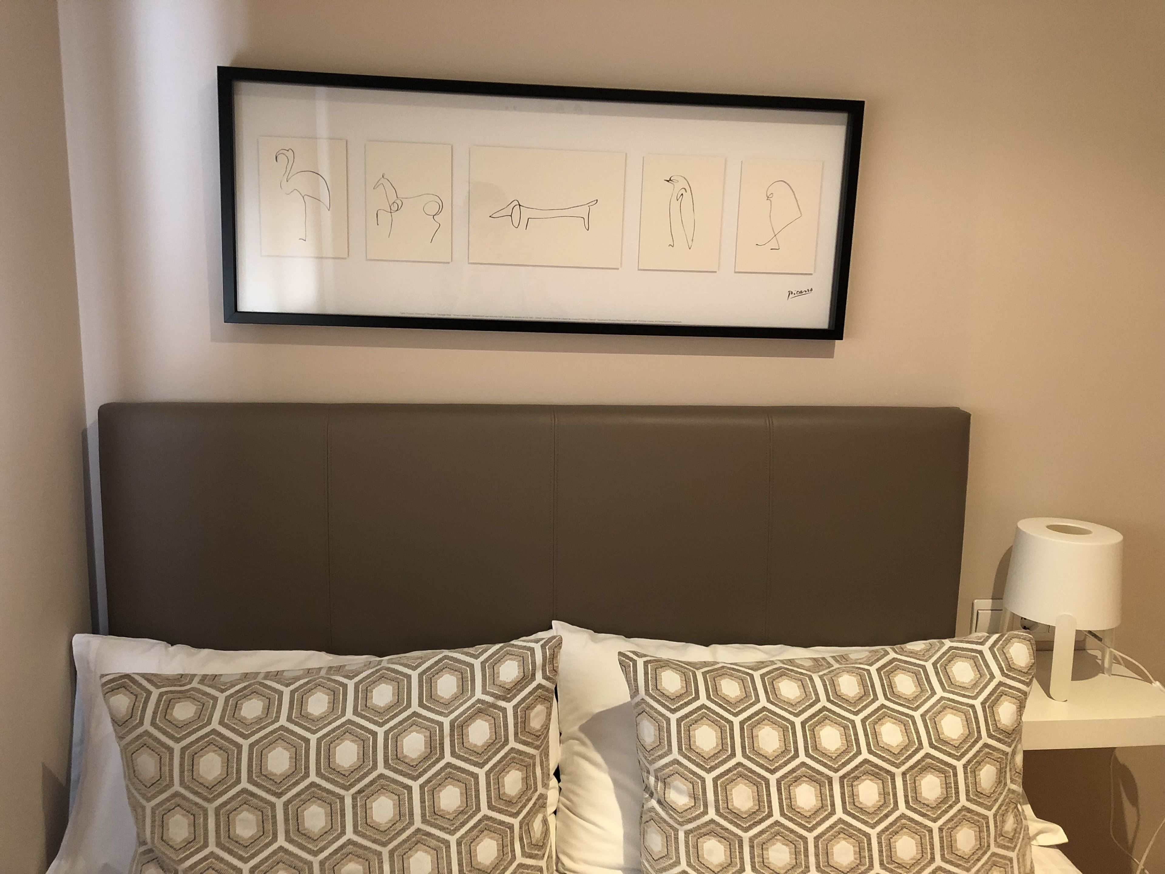 Ein Schlafzimmer mit einem Kopfteil, einem gerahmten Bild mit Tierkonturen, gemusterten Kissen und einer Nachttischlampe.
