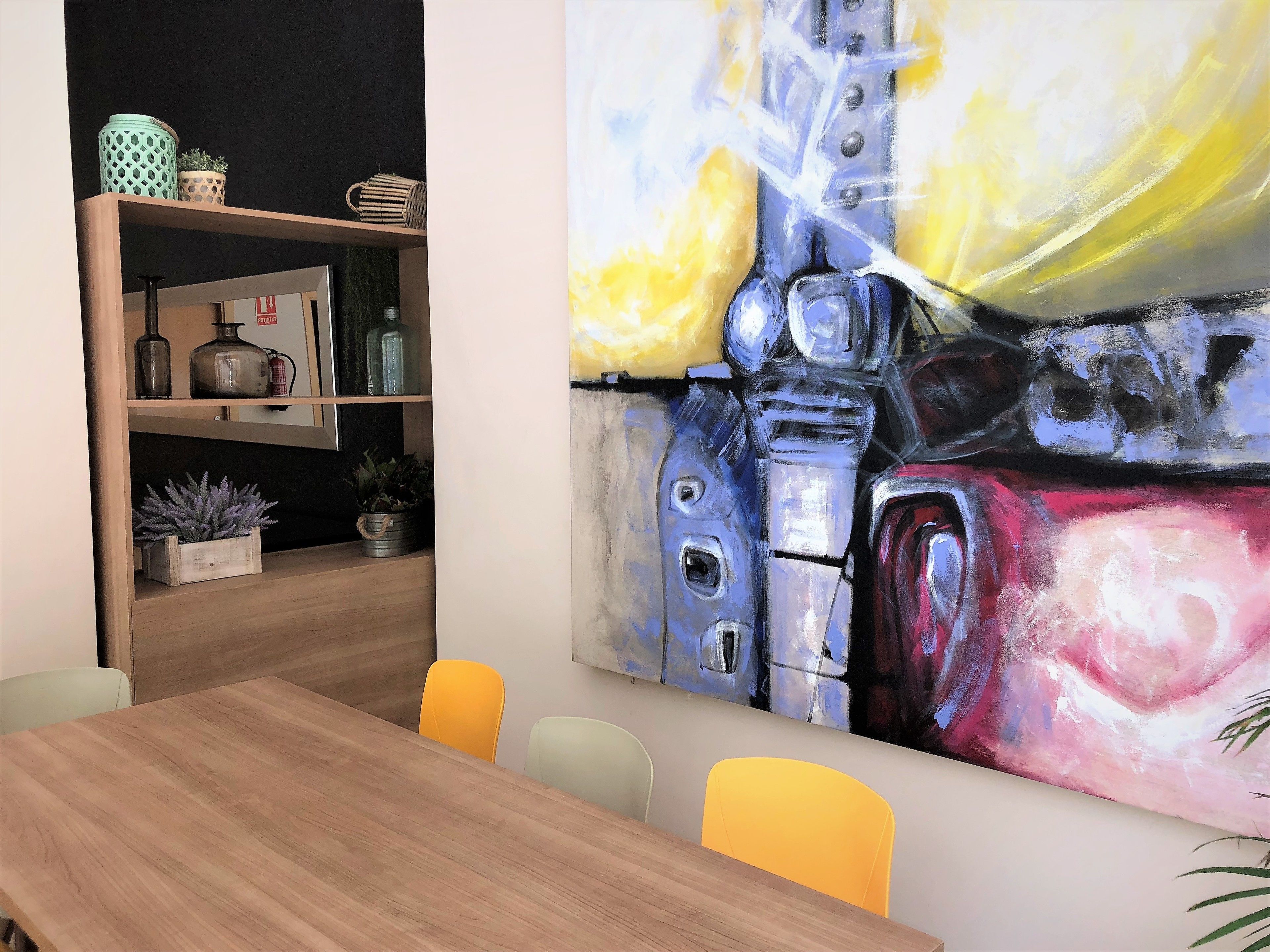 A modern dining area with a wooden table and chairs, a bookshelf with decorative items, and a large abstract painting on the wall.