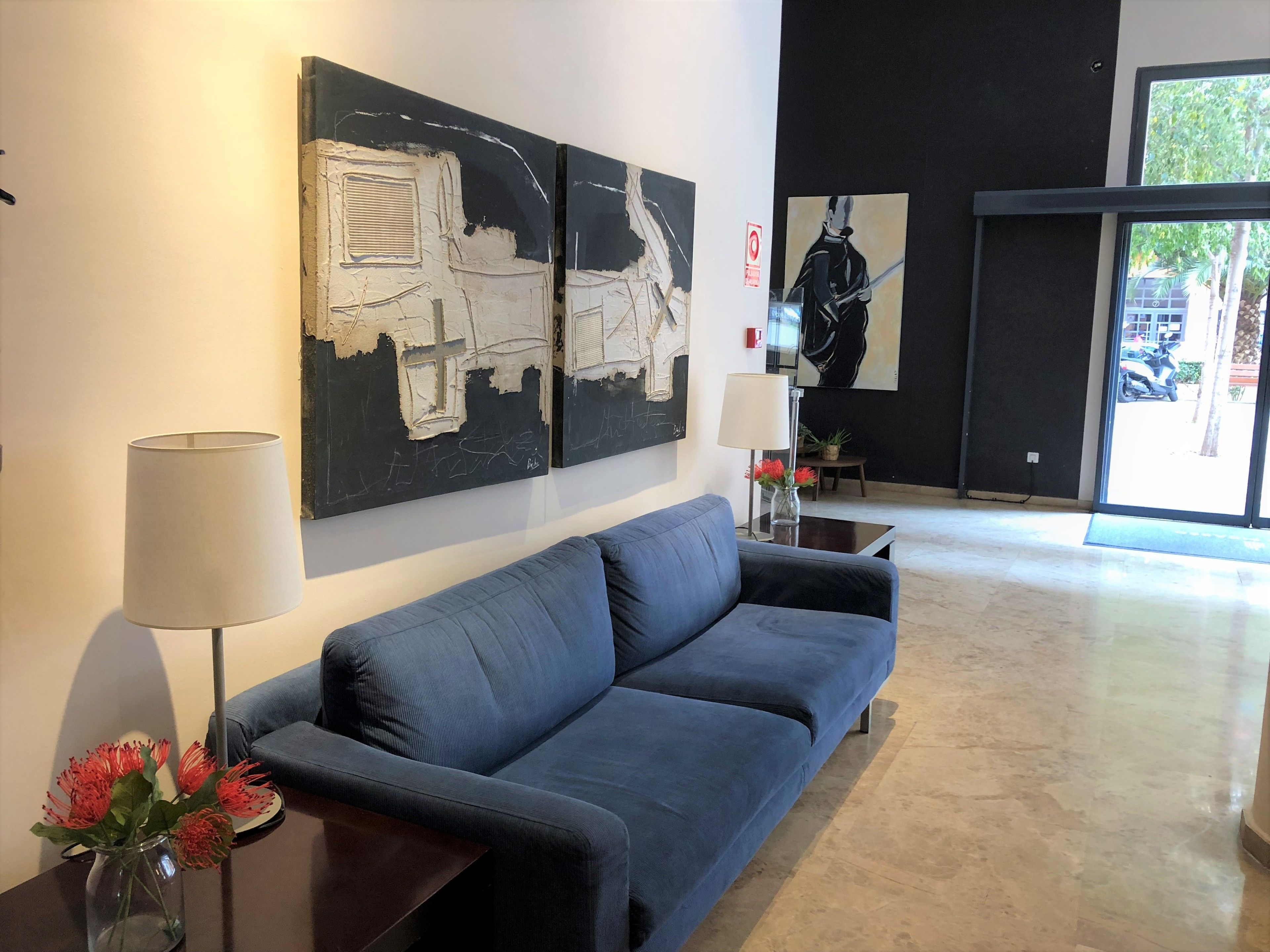 A modern lobby with a blue sofa, abstract wall art, a vase with flowers, and a lamp.