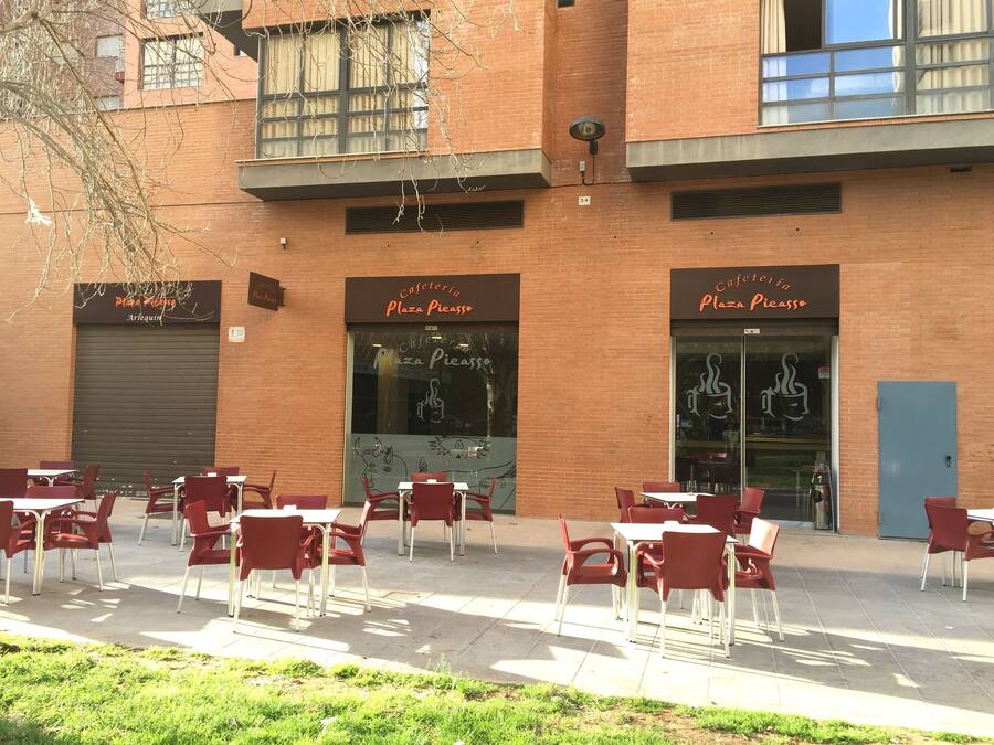 Un café al aire libre con sillas y mesas rojas, un edificio de ladrillo y un amplio ventanal con un mural.