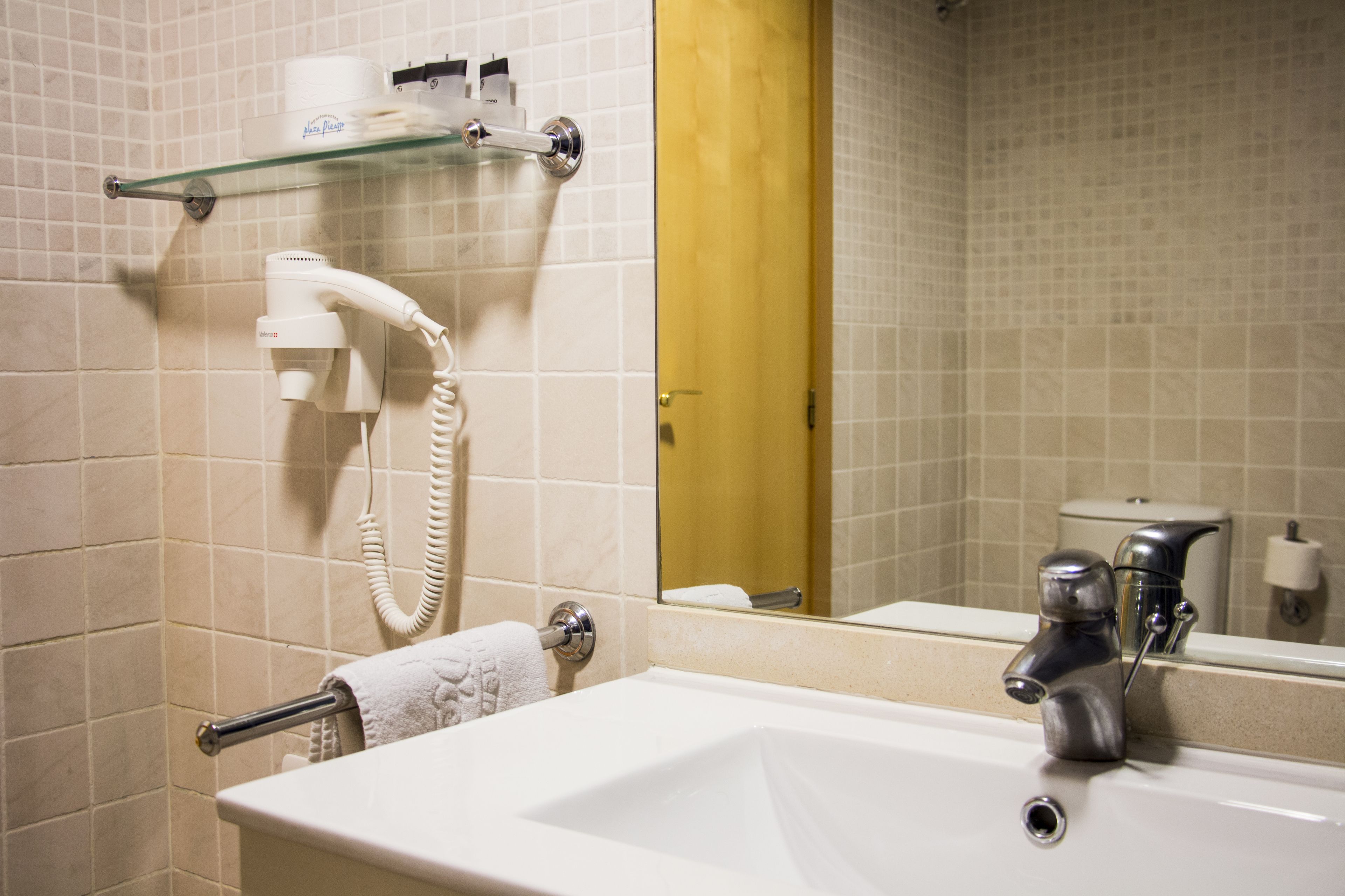 A bathroom with a white sink, a glass shelf with toiletries, a hairdryer, and a toilet visible in the reflection.