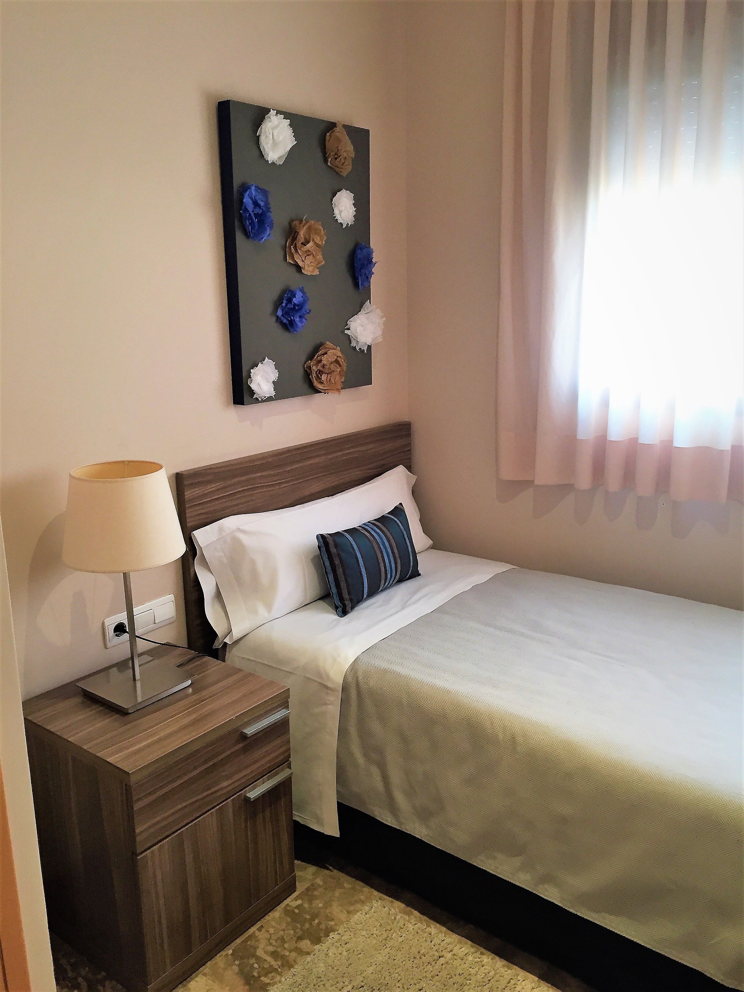 Een net opgemaakt bed met een witte deken en een blauw gestreept kussen, een nachtkastje met een lamp, en een kunstwerk aan de muur erboven.