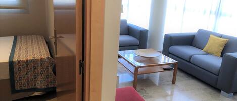 Una sala de estar con un sofá, una mesa de centro, un reposapiés rojo y una colcha estampada.