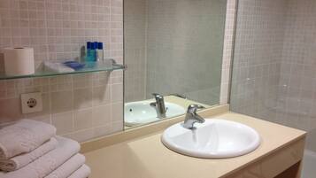 Un baño con un lavamanos blanco, un estante de vidrio con artículos de aseo y una pila de toallas blancas.