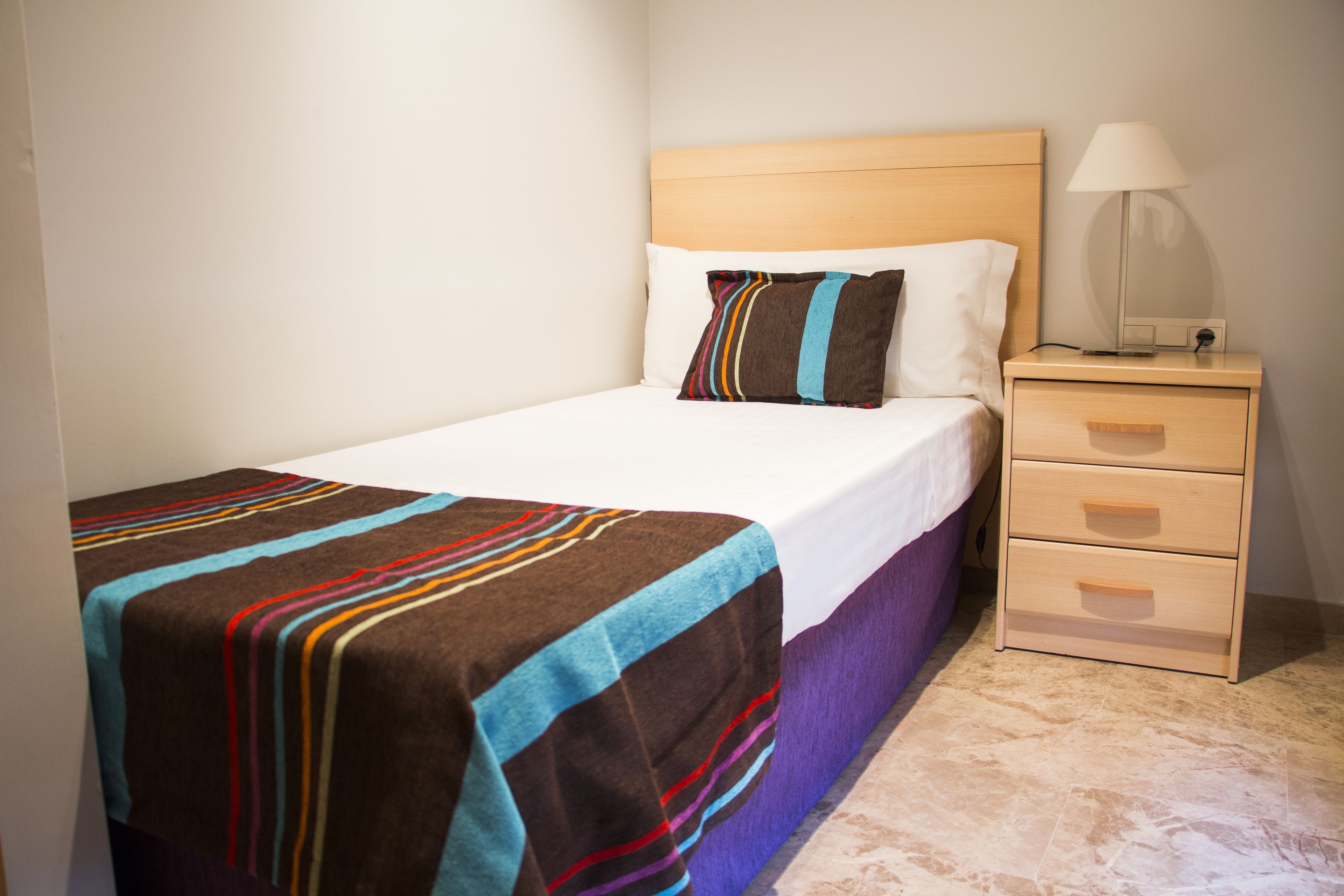 A neatly made bed with a striped blanket, a bedside lamp, and a wooden nightstand with drawers.
