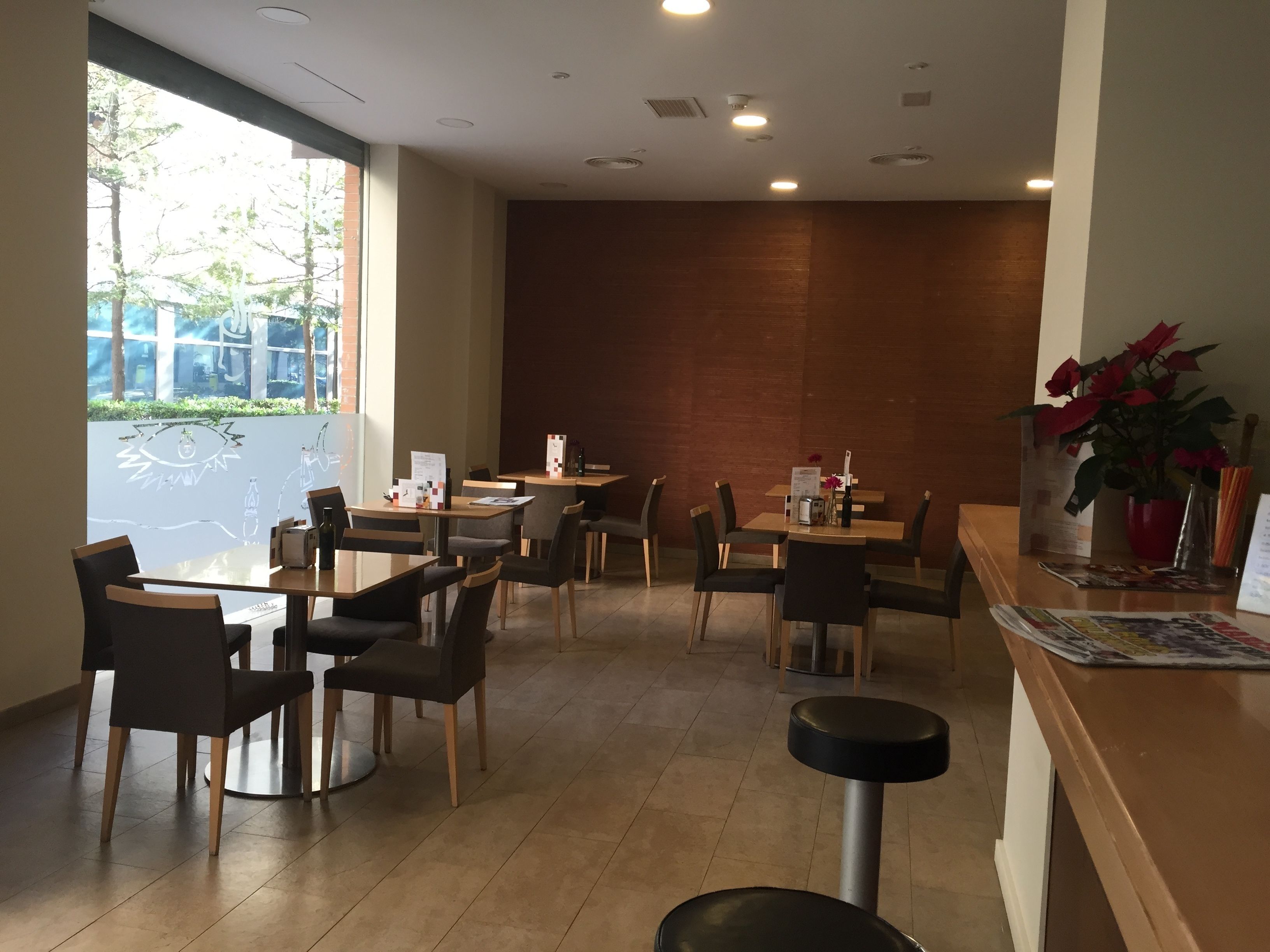 A restaurant with multiple tables and chairs, a counter with a menu, and a large window with a view of trees and buildings.