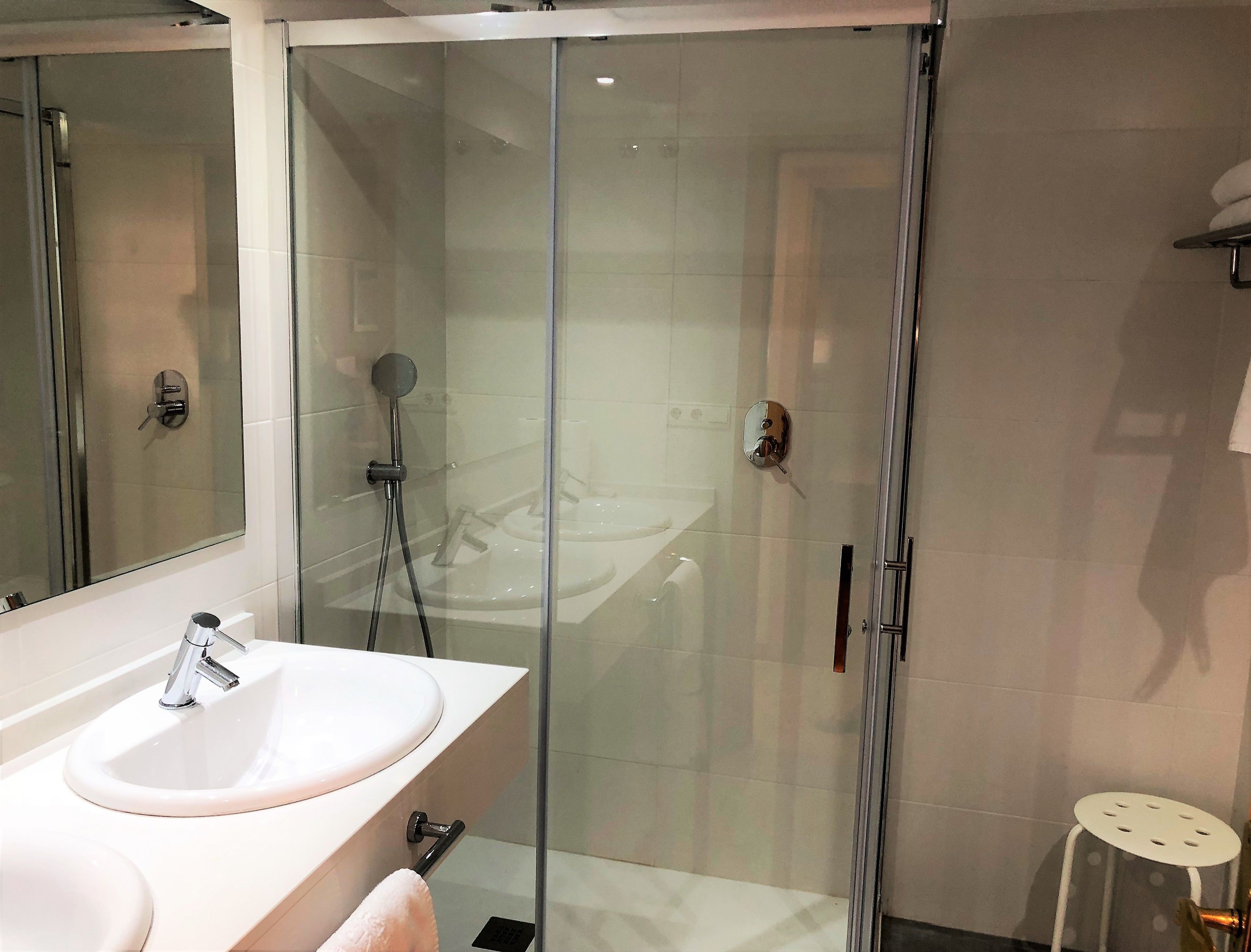 Een moderne badkamer met een glazen douchecabine, een witte wastafel met spiegel, en een klein krukje.
