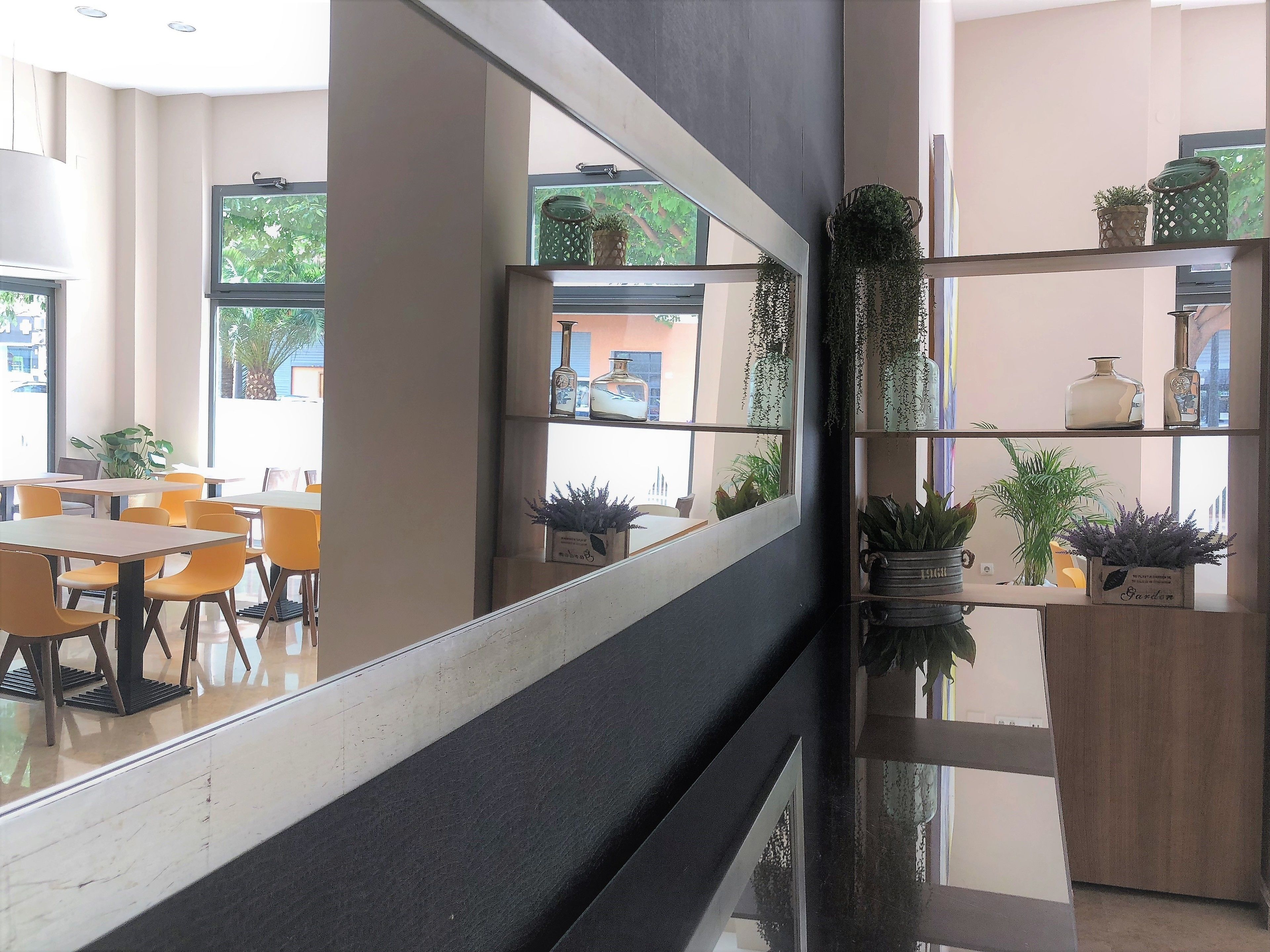 A modern interior with a long reflective corridor, seating area with tables and chairs, and shelves with potted plants and decorative items.