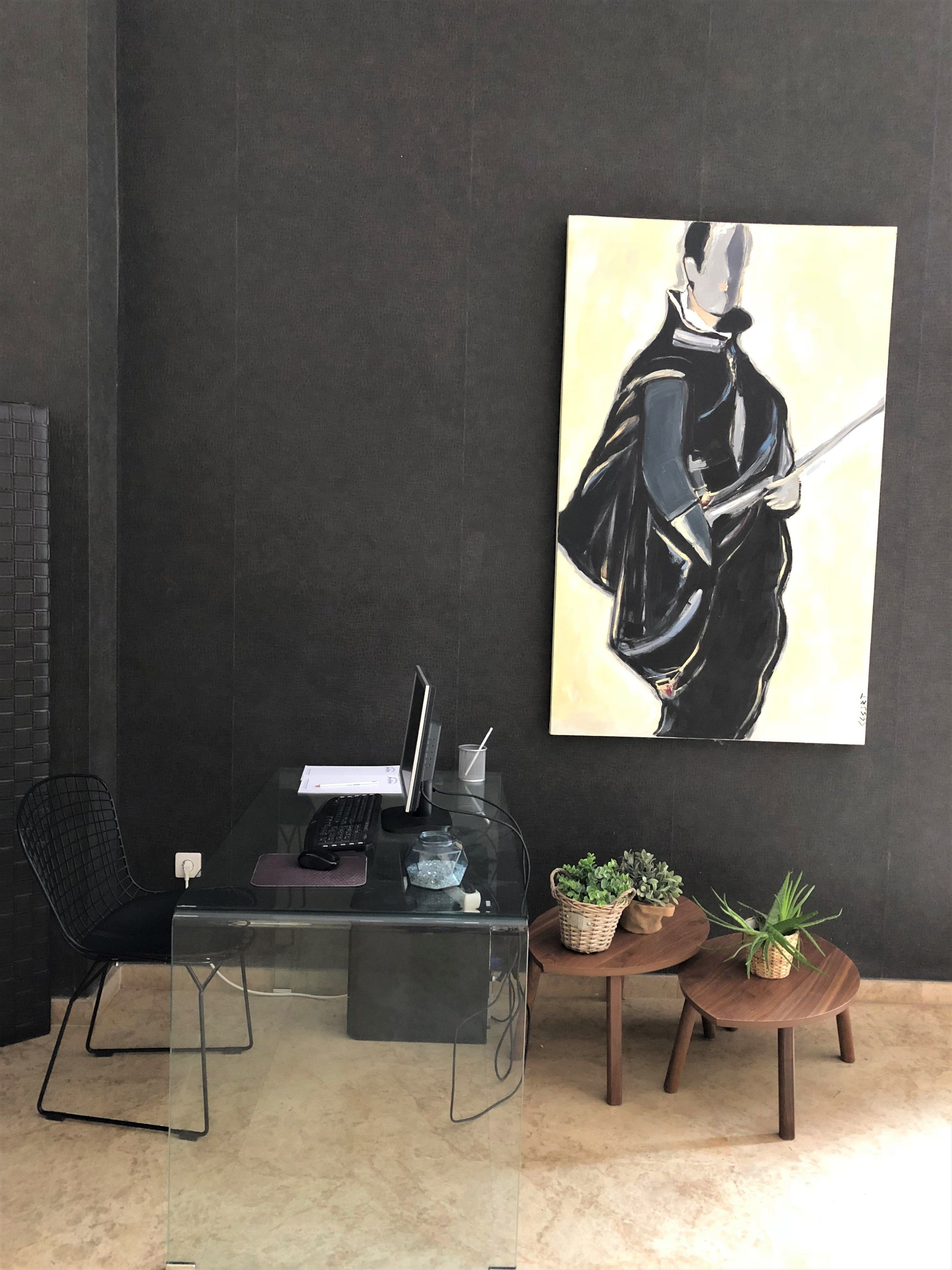 A modern office space with a glass desk, a computer, and a black chair. A large painting of a figure in black is on the wall. There are two small wooden tables with plants.