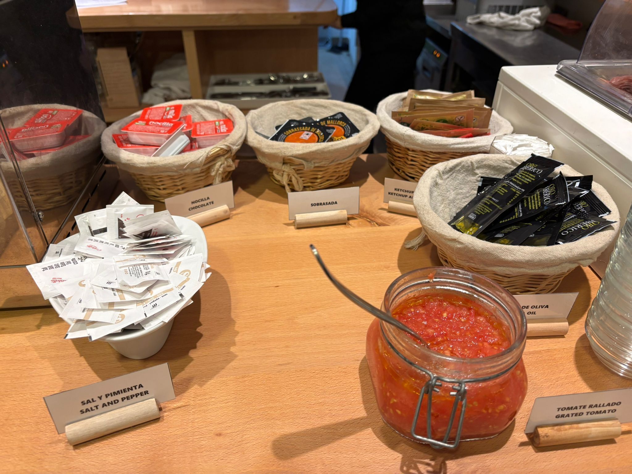 Taula de buffet amb diversos plats i condiments, incloent cistells amb noms dels aliments.
