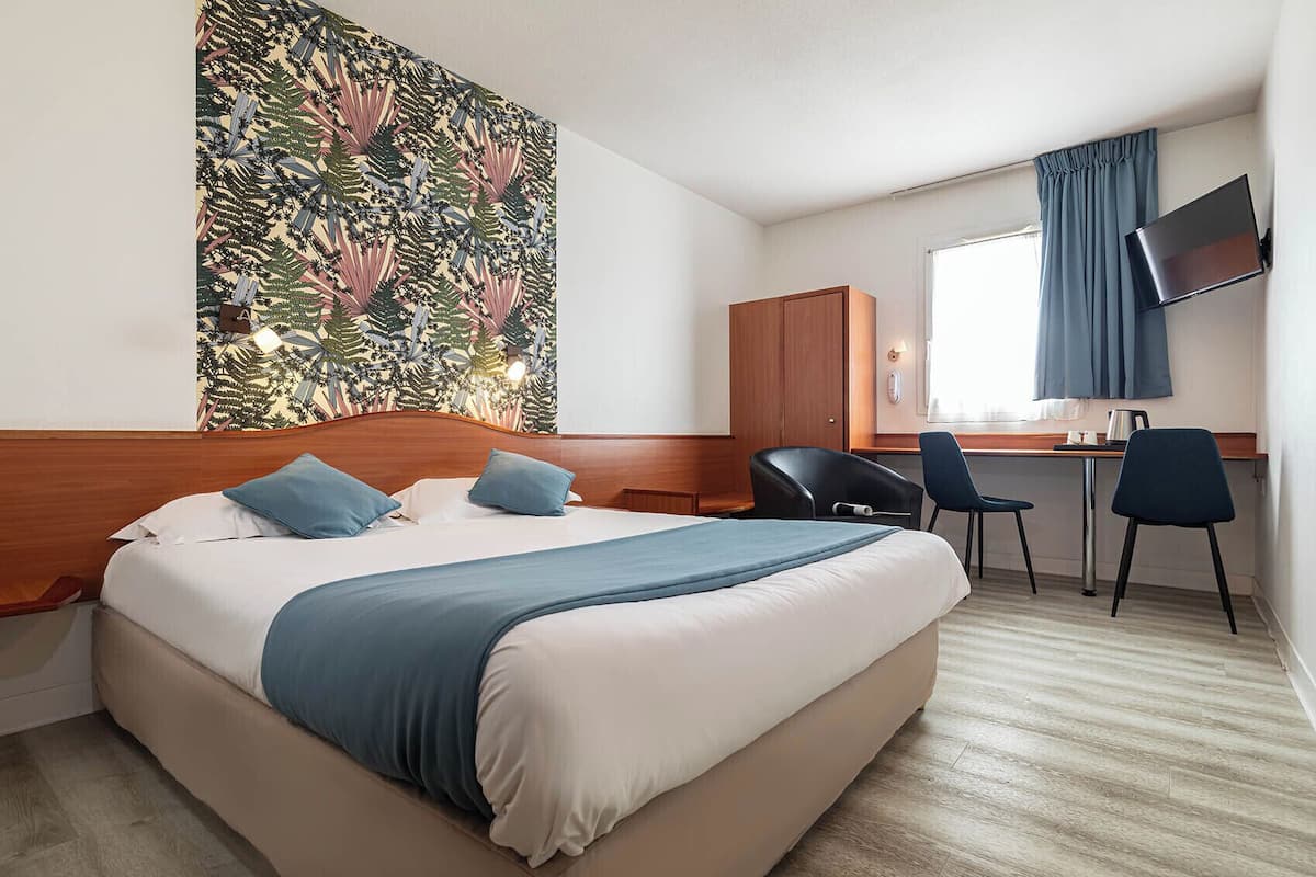 A hotel room with a large bed, a desk, two chairs, a TV, and a wall with a tropical leaf pattern.
