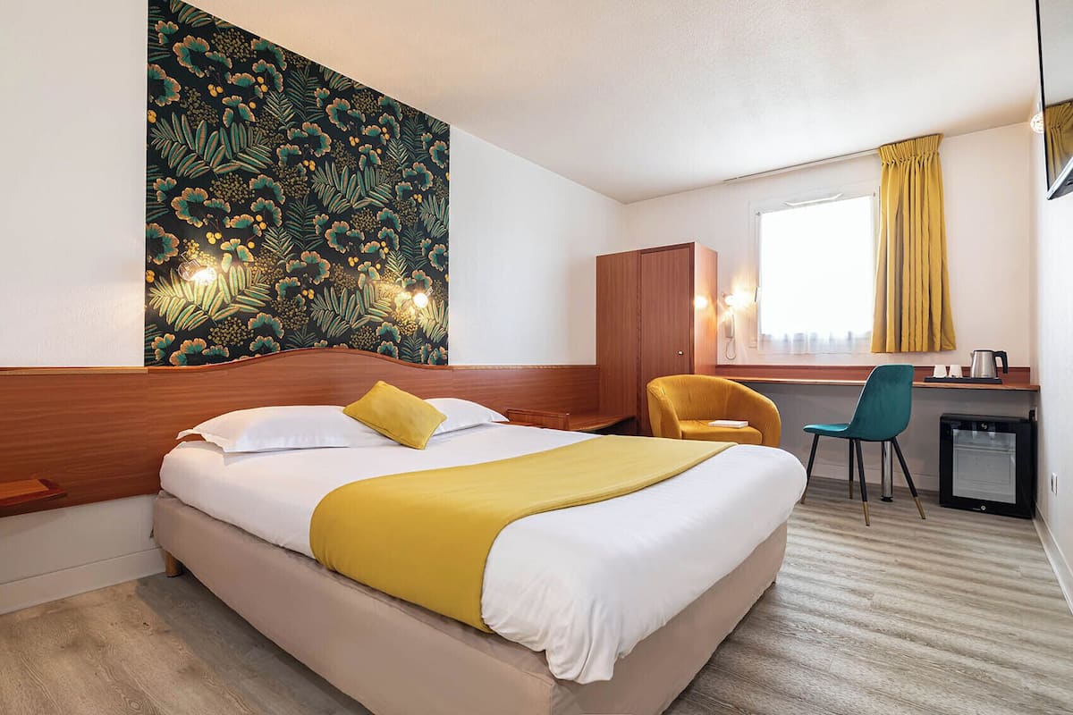 A hotel room with a bed, a desk, a chair, a small kitchenette, and a wall with a floral pattern.