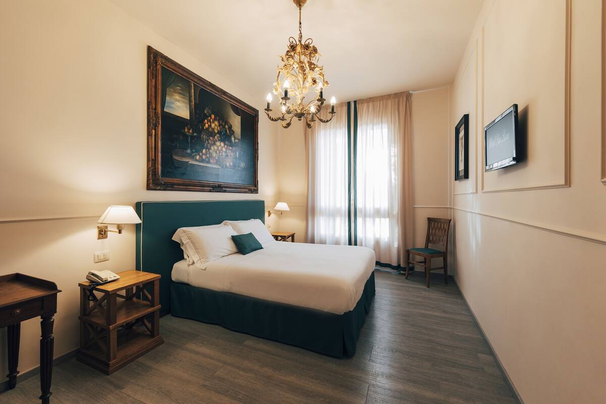 A hotel room with a large bed, a chandelier, a painting, a television, and a chair.