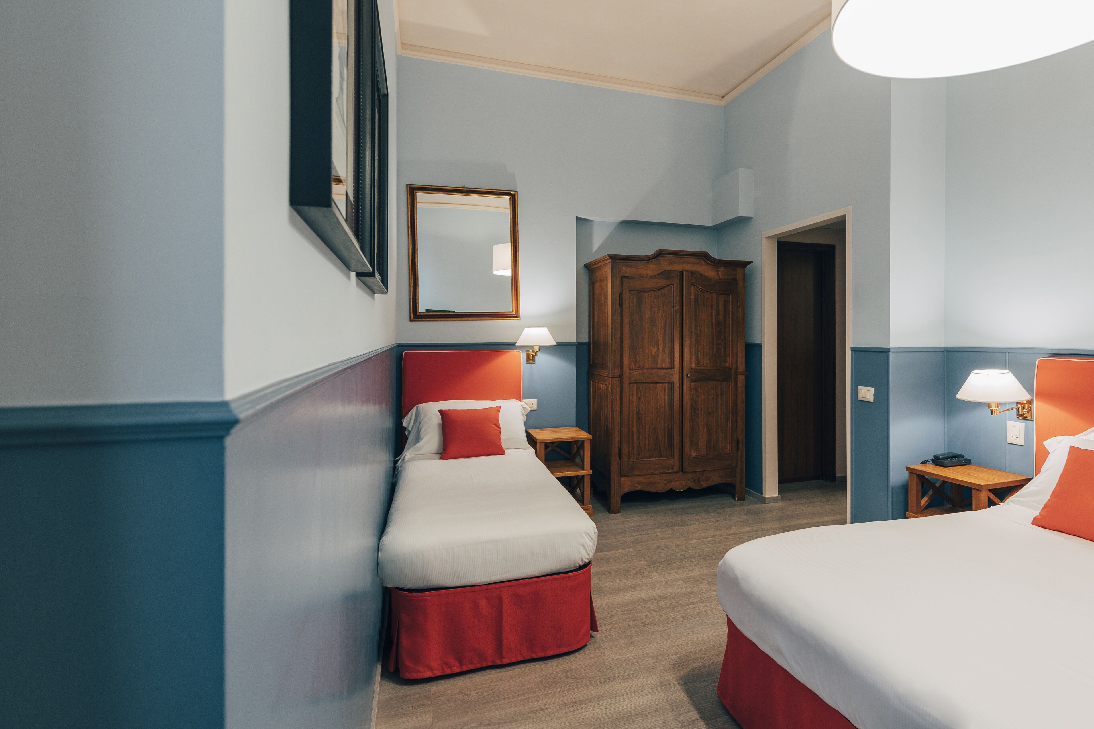 Ein Hotelzimmer mit zwei Betten, einem Holzschrank, einem Spiegel und einem kleinen Tisch mit einer Lampe.