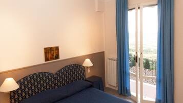 Ein Hotelzimmer mit einem großen Bett, zwei Nachttischlampen, einem Kopfende mit Muster sowie einem kleinen Gemälde an der Wand und einem Balkon mit blauen Vorhängen.