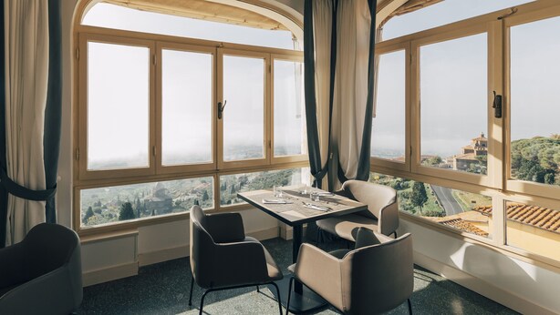 A dining area with a table and chairs, large windows with curtains, and a view of a landscape. - Hotel San Luca (Cortona)