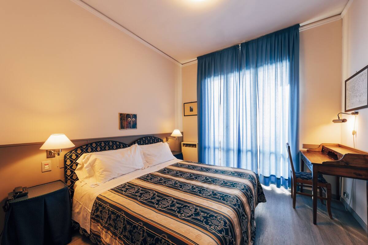 A hotel room with a bed, a desk, and a chair. There are two lamps on either side of the bed.