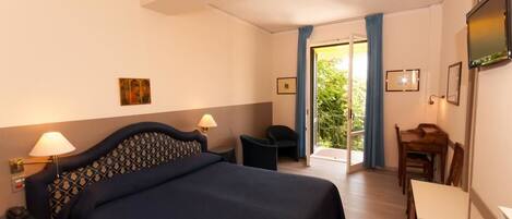 Habitación de hotel con cama, escritorio y silla. Cuenta con una ventana con cortinas, un televisor montado en la pared y una puerta que da a un espacio exterior.