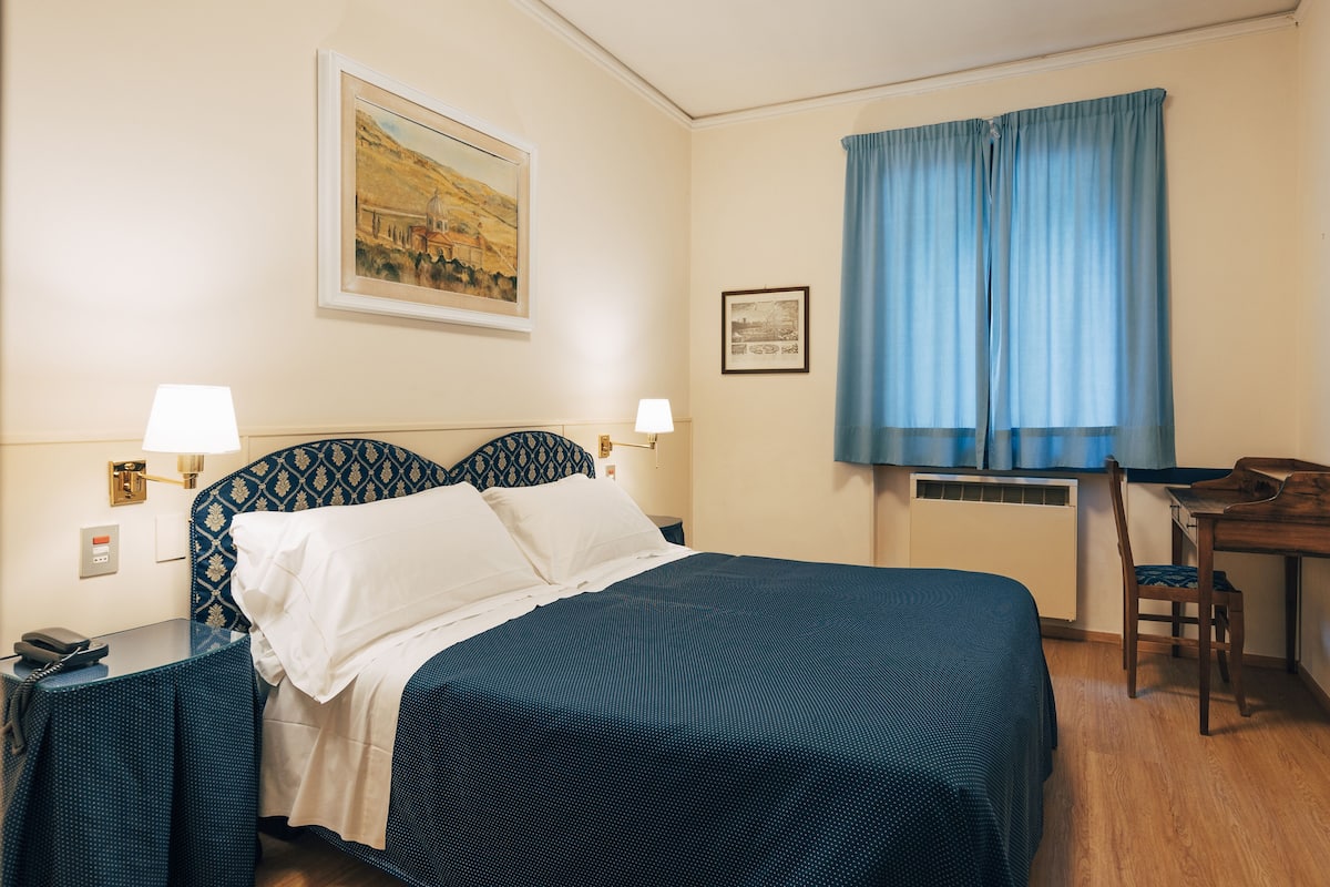 A hotel room with a bed, two bedside lamps, a desk with a chair, a telephone, and a window with curtains.