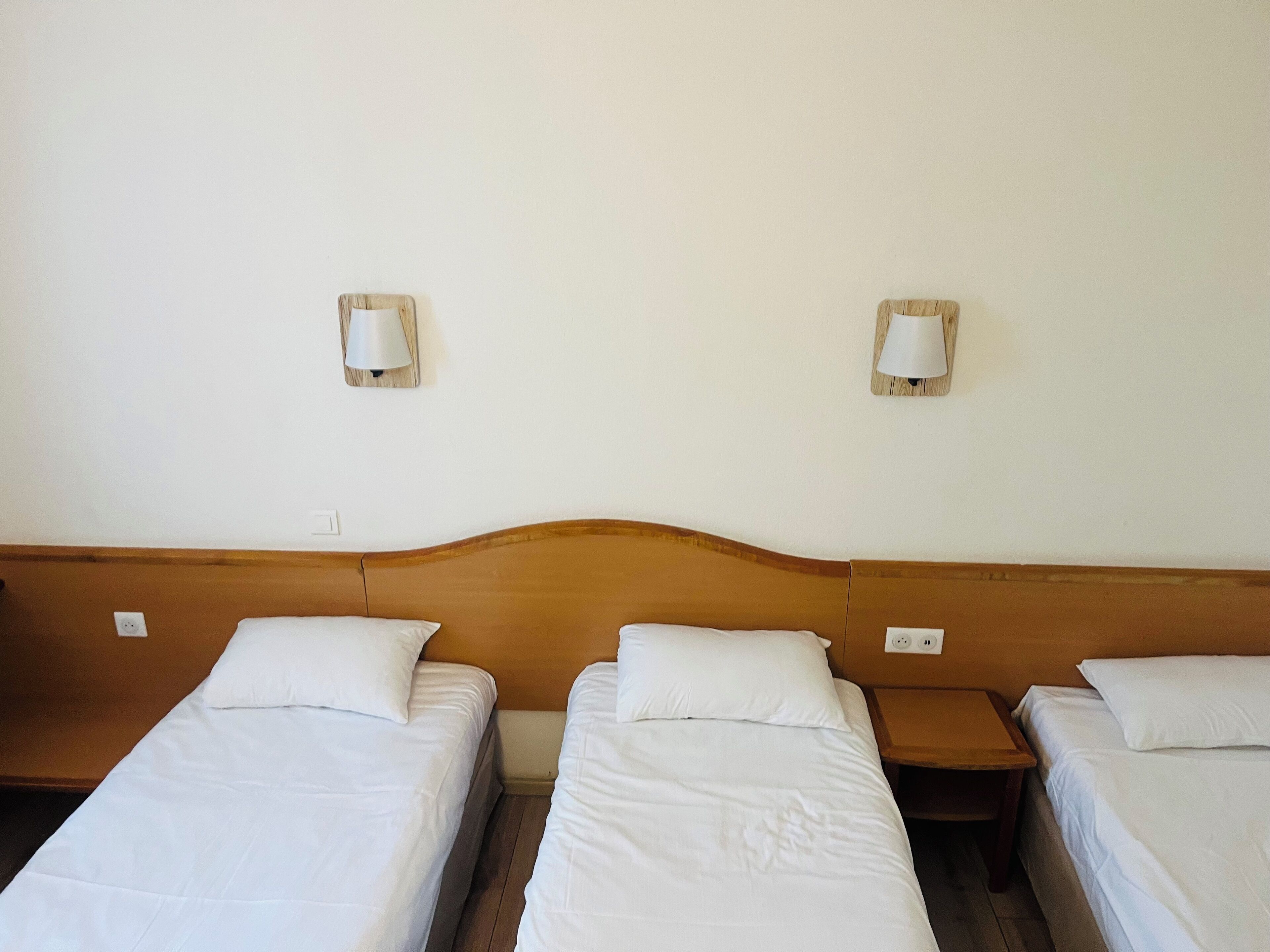 Habitación de hotel con tres camas individuales, cada una con ropa blanca y cabeceros de madera. Dos lámparas de pared a cada lado de las camas.