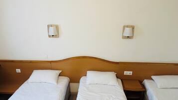Une chambre d’hôtel avec trois lits simples, chacun doté de draps blancs et d’une tête de lit en bois. Deux lampes murales sont fixées de chaque côté des lits.