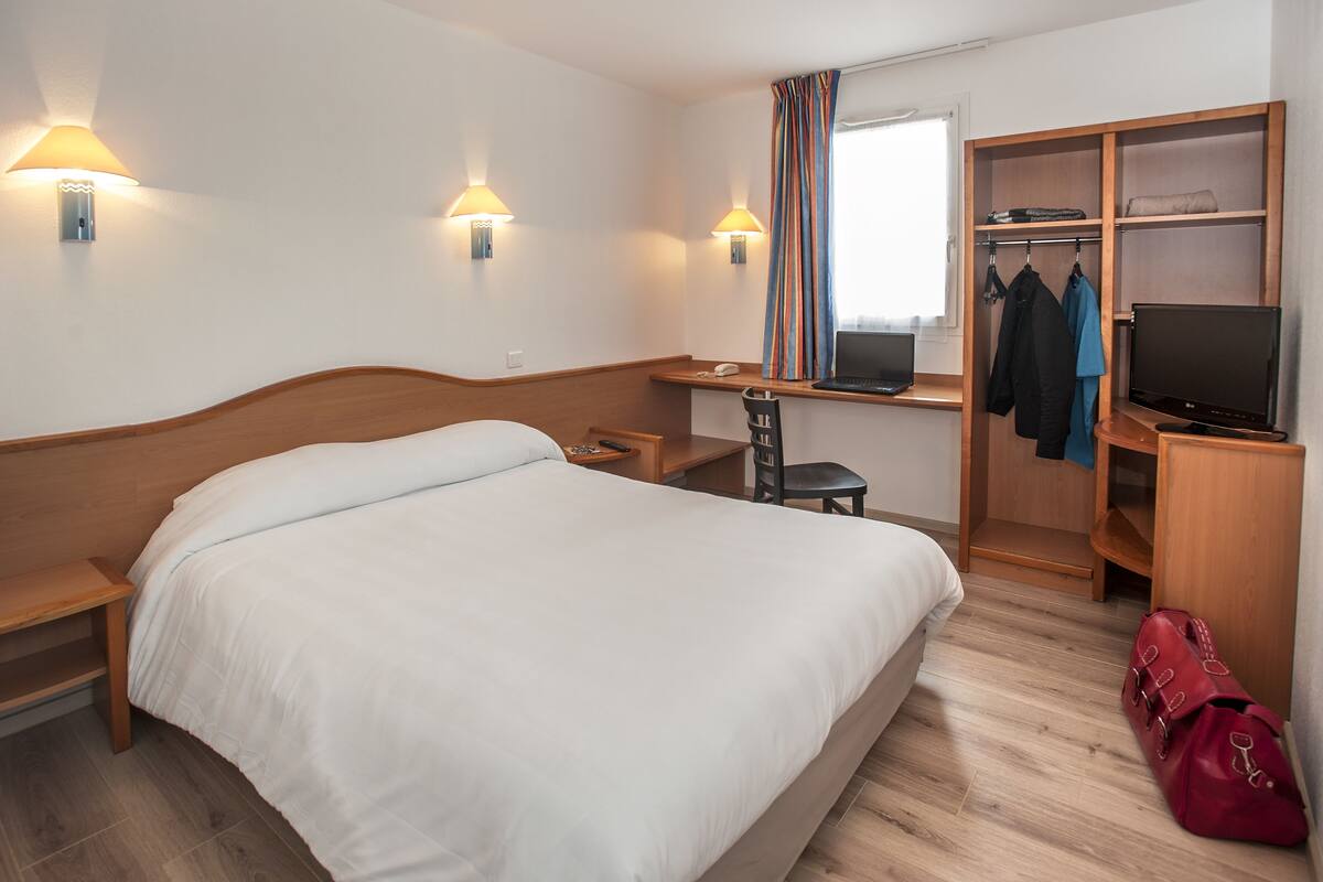 Une chambre d’hôtel avec un lit, un bureau, une chaise, une télévision, une armoire et un sac rouge.