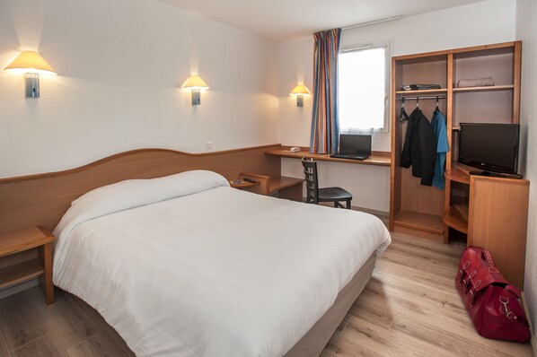 A hotel room with a bed, desk, chair, TV, wardrobe, and a red bag.