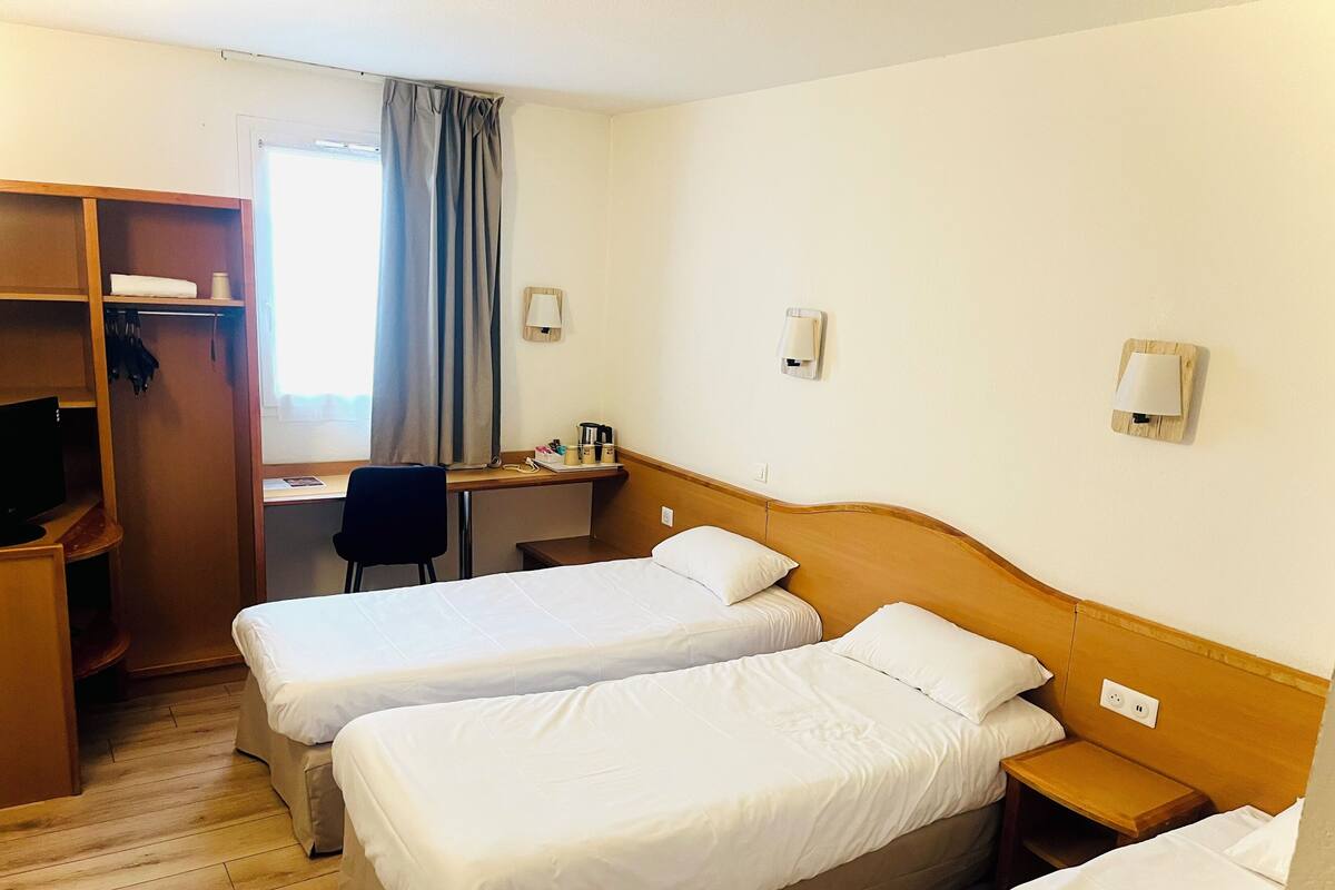 Une chambre d’hôtel avec deux lits simples, un bureau avec une chaise, une télévision et une armoire.