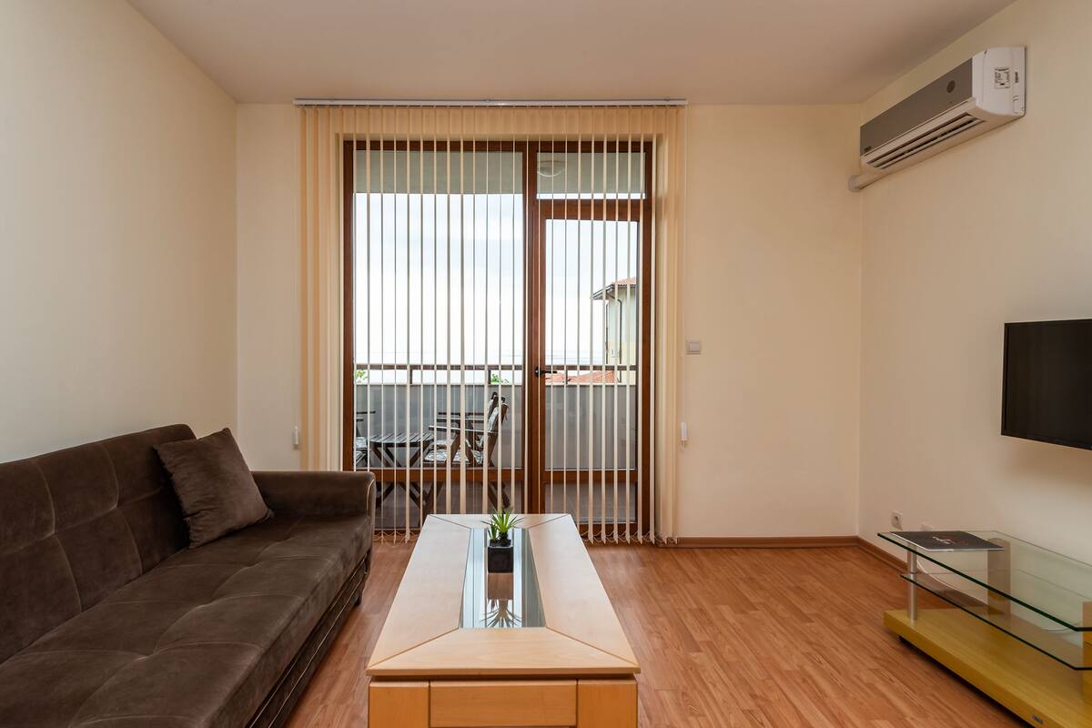 A modern living room with a brown sofa, a glass coffee table with a plant, wooden flooring, a sliding glass door with blinds, a balcony, and a mounted TV.