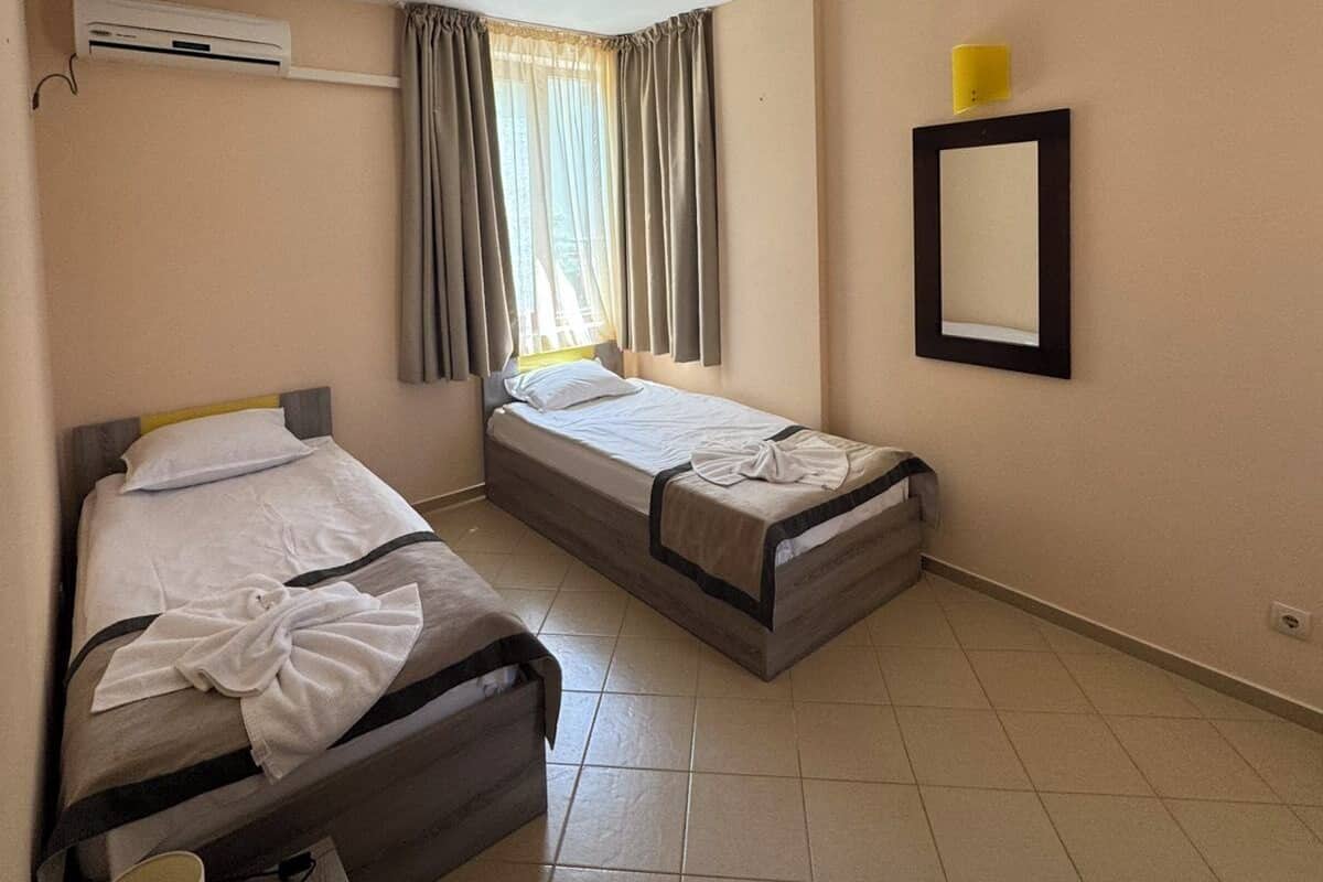 A hotel room with two beds, a window with curtains, an air conditioning unit, a bedside table with a lamp, and a wall-mounted mirror.