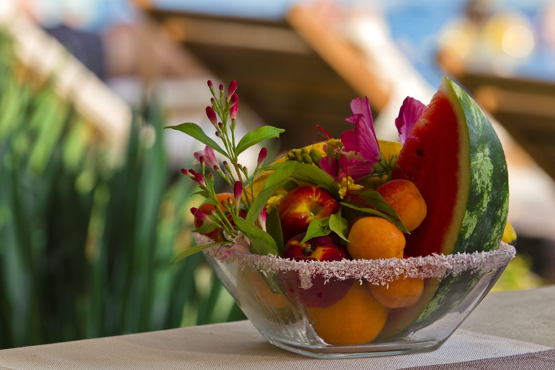A clear glass bowl containing a variety of fruits and flowers, placed on a table with a blurred outdoor background.