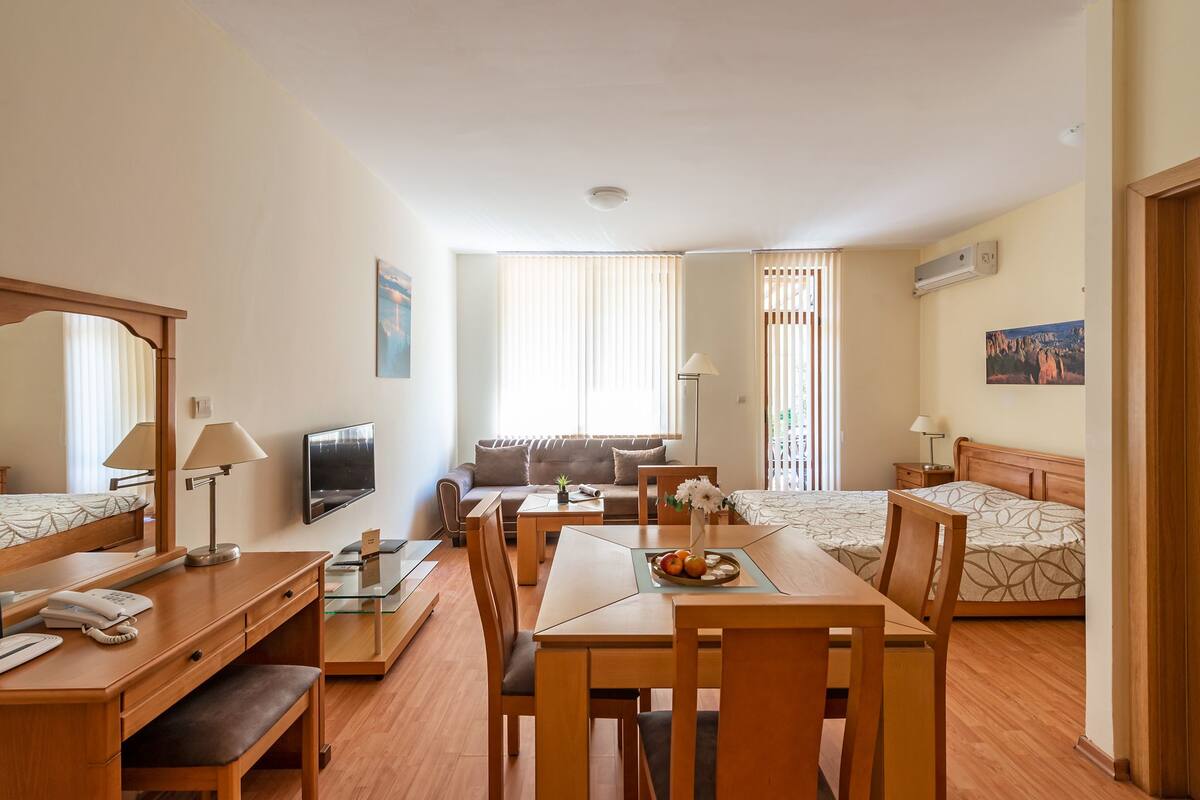 A hotel room with a dining area, a sofa, two beds, a TV, and a window with curtains.