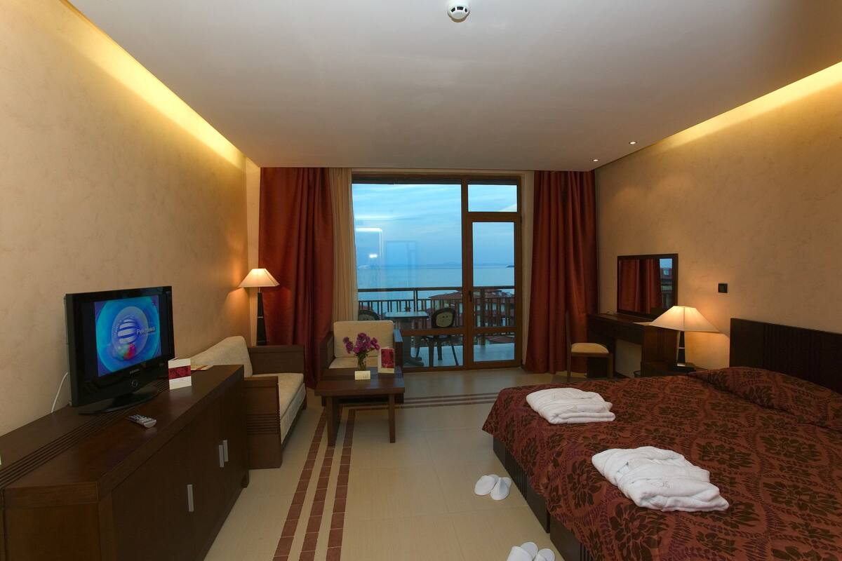 A hotel room with a bed, a desk, a television, a balcony with a view of the sea, and a window with curtains.