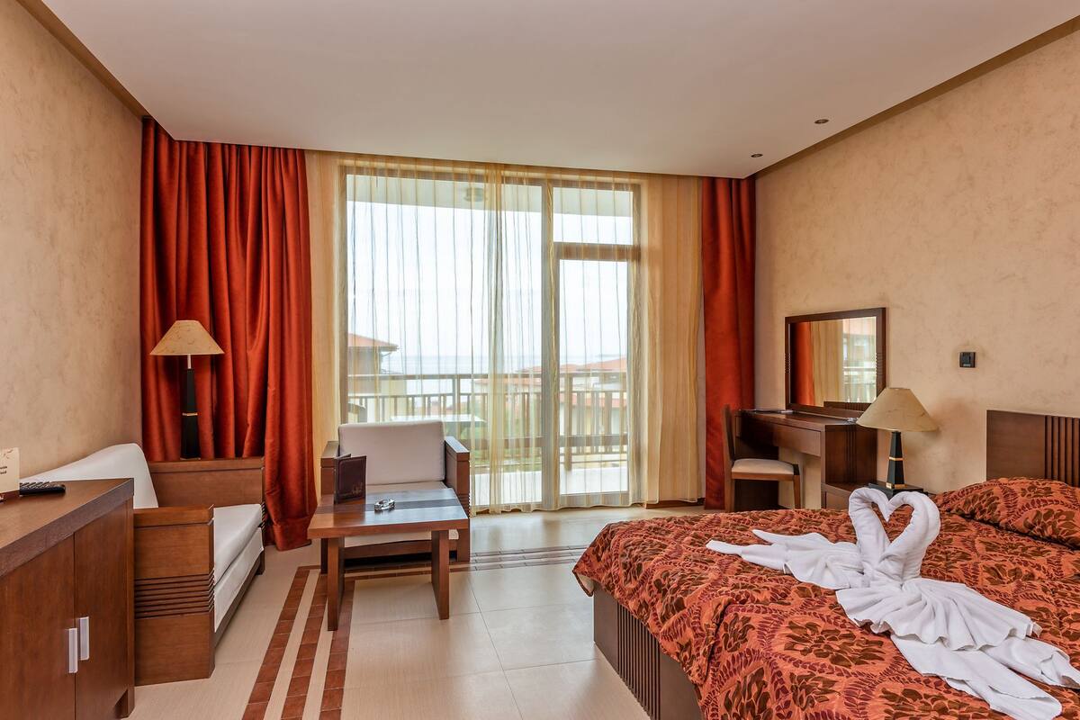 A hotel room with a bed, a desk, a chair, a small table, and a balcony with curtains.