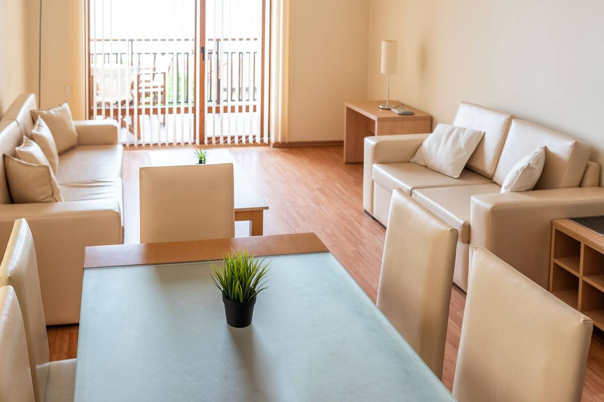 A modern living room with a sofa set, a coffee table with a plant, and a view of the outdoors through large windows.