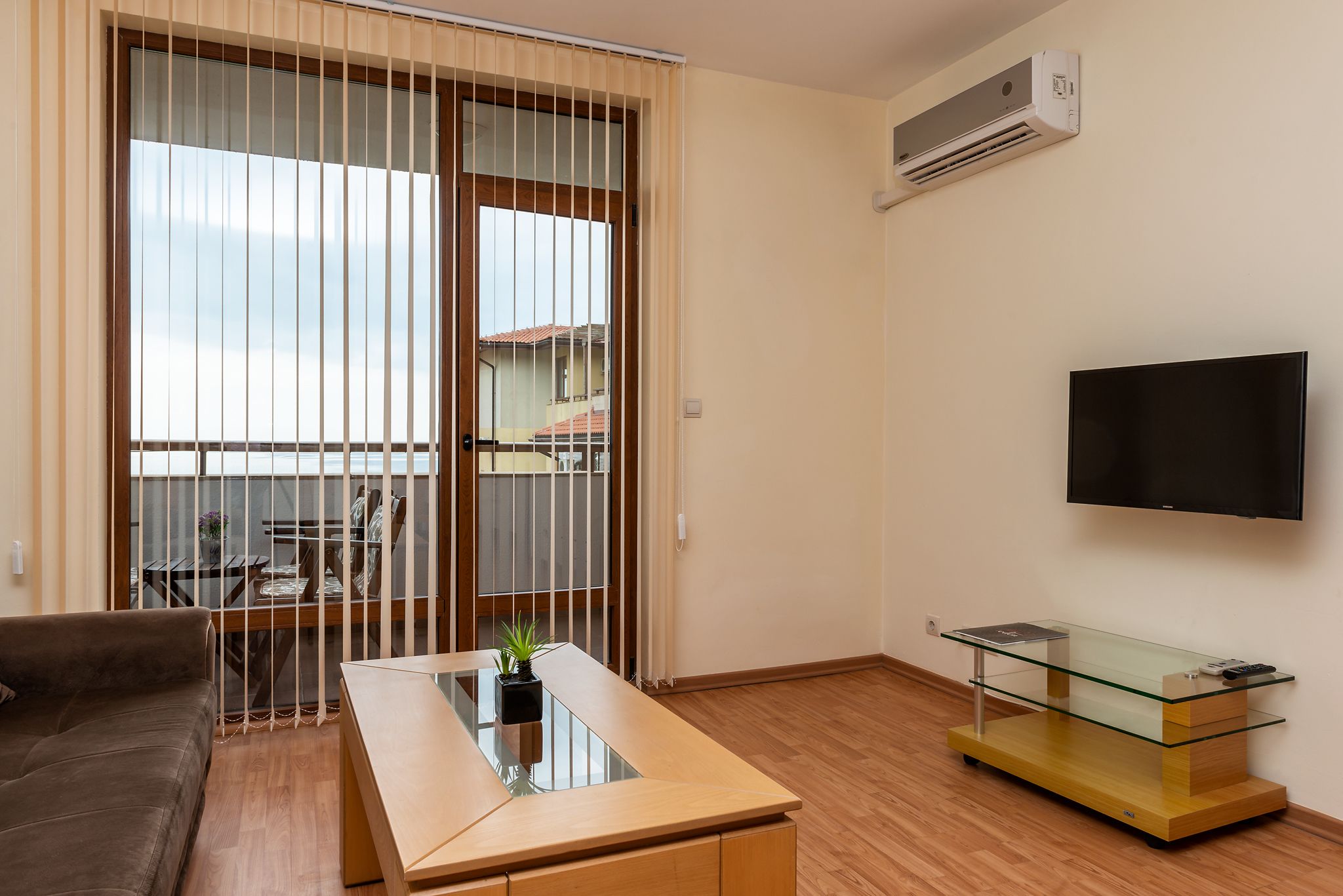Una sala de estar moderna con una mesa de centro de vidrio, un sofá, un televisor sobre un mueble bajo y un ventanal corredizo que da a un balcón.