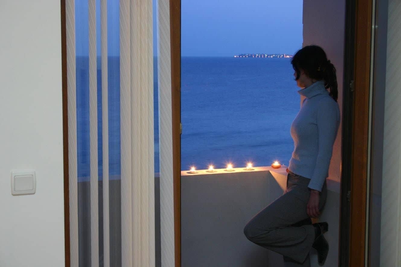 Una mujer apoyada en la barandilla de un balcón, con vista al mar y un muelle distante.