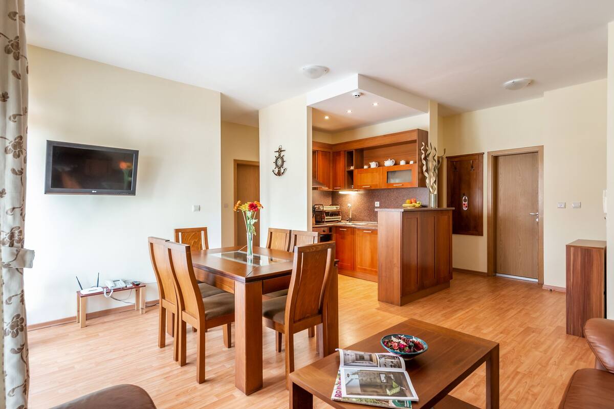 A modern living room with a wooden dining table, chairs, and a TV mounted on the wall. A kitchen with wooden cabinets and a central island is visible in the background.