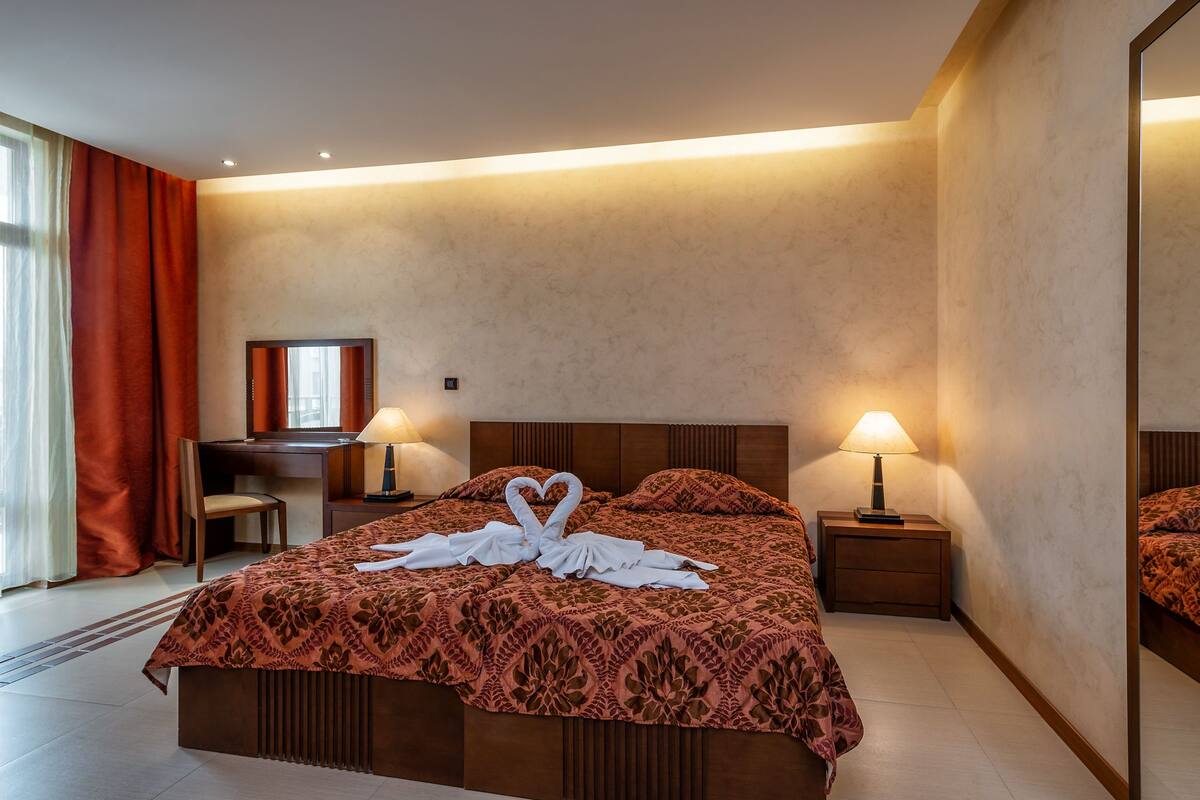 A hotel room with a bed, two bedside lamps, a desk with a chair, and a mirror on the wall.