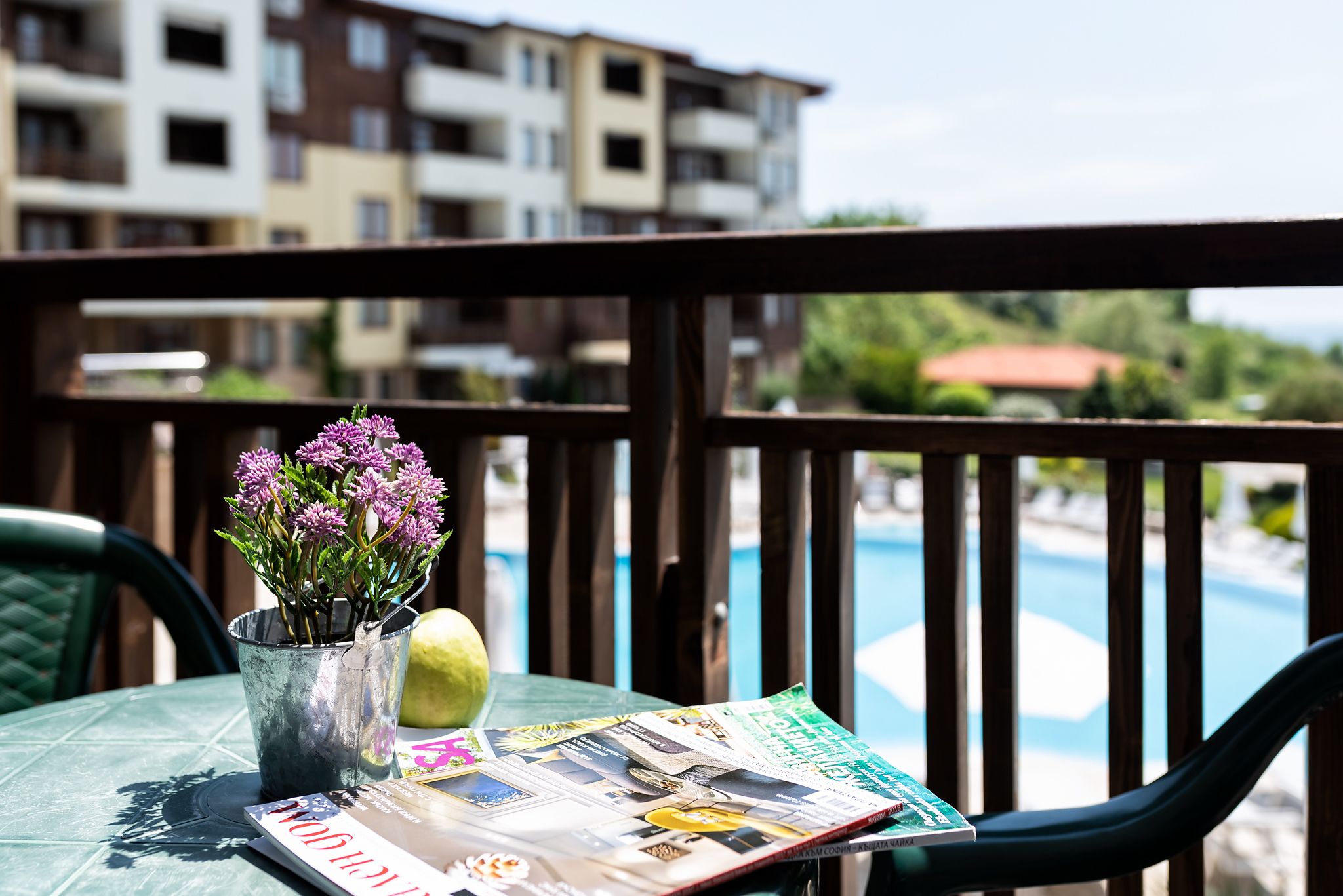 A table with a magazine, a potted plant, and a lemon on a balcony overlooking a swimming pool.